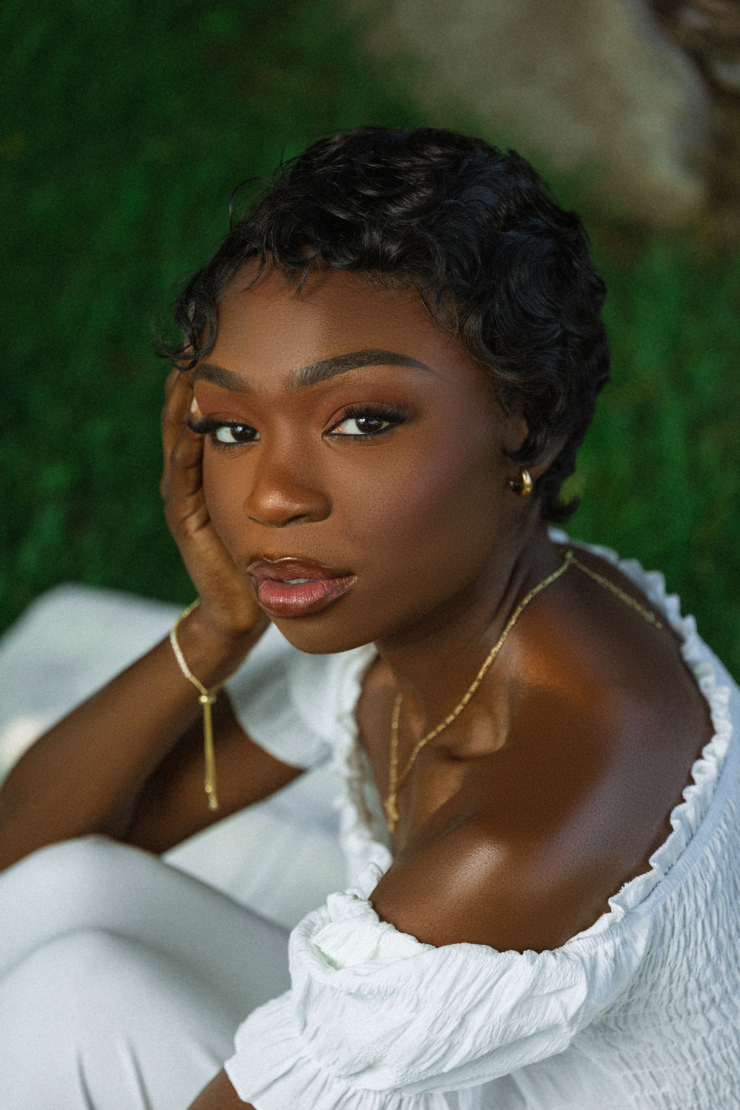 Beautiful portrait in soft lighting featuring elegant white off-shoulder top and delicate gold earrings against lush green background.