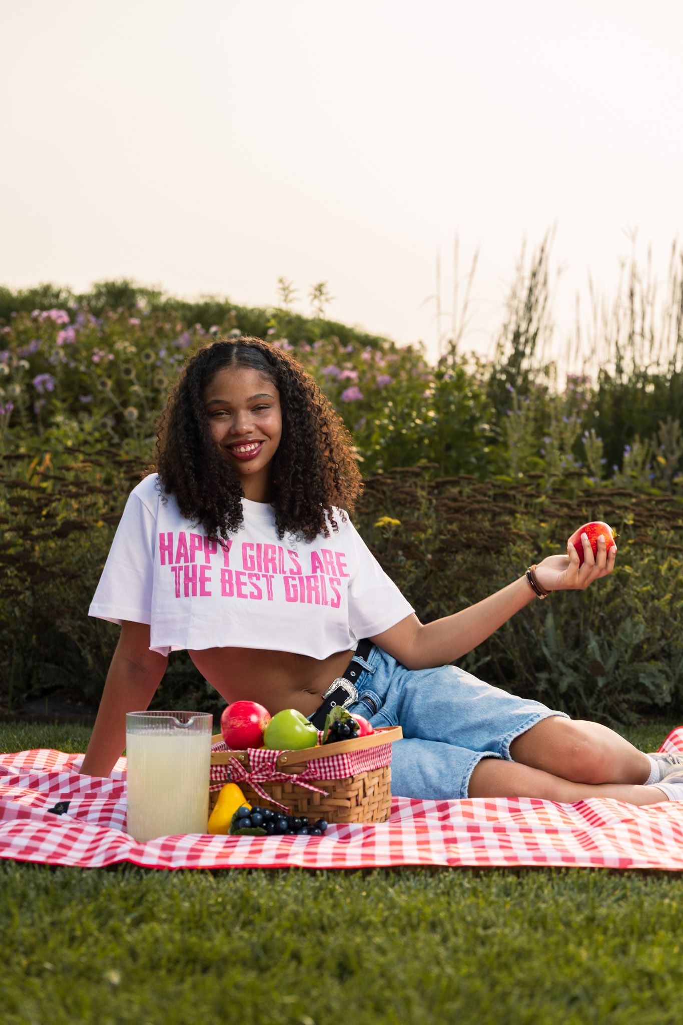Woman enjoying a sunny picnic outdoors on red checkered blanket with fresh fruits and snacks.
