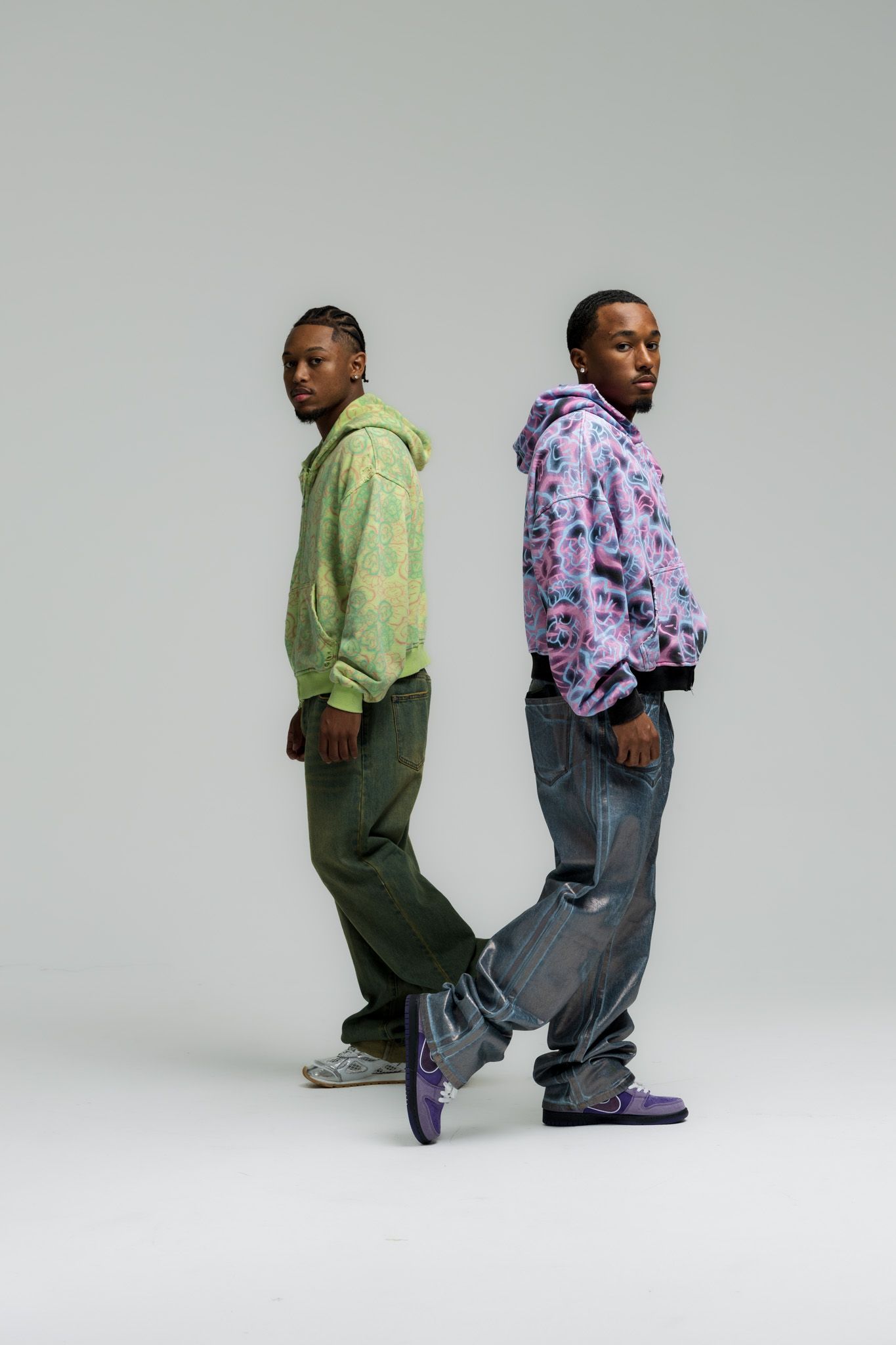 Two people stand back-to-back wearing colorful hoodies and baggy pants in a minimalist studio setting.
