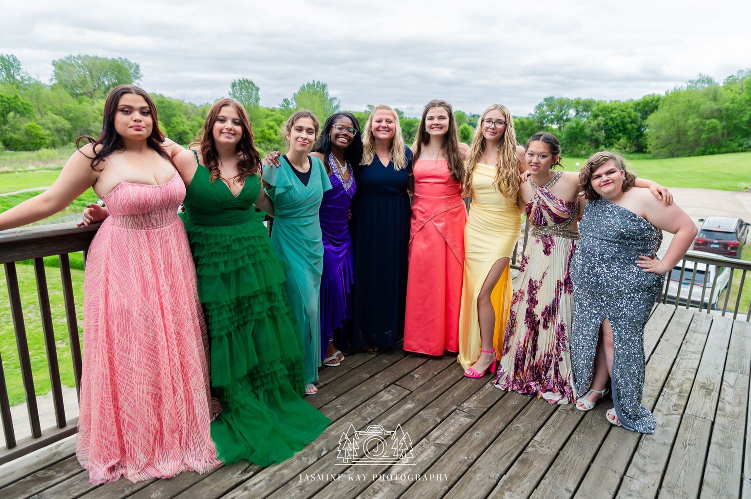 A group wearing colorful formal dresses pose together on a wooden deck during a special occasion.