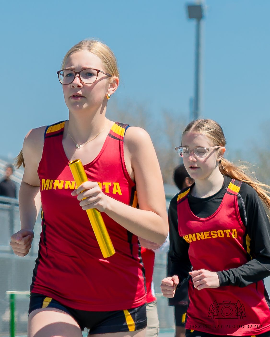 Two athletes in red Minnesota jerseys running during a track competition against a blue sky background.
