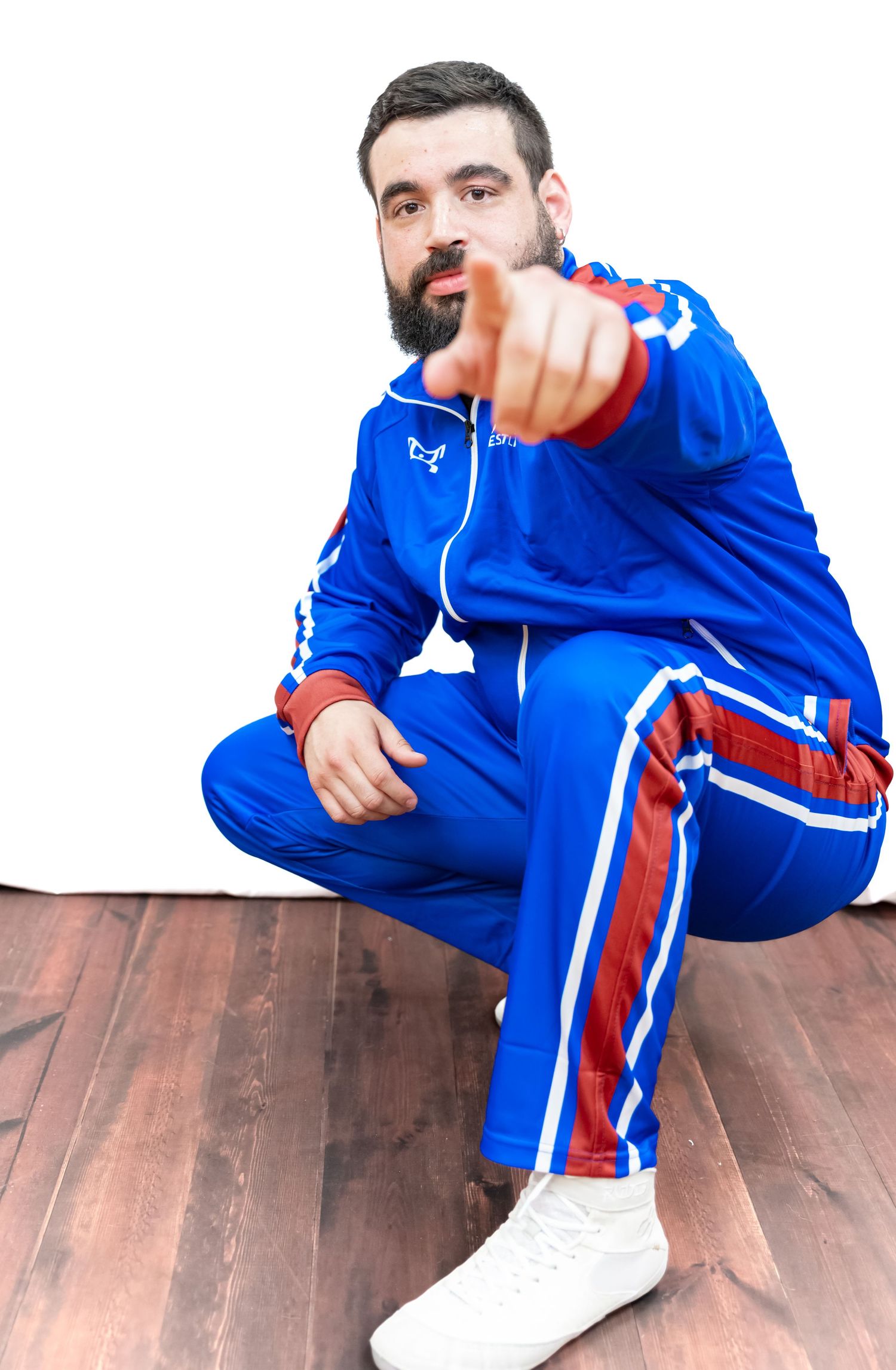 Athletic figure in bright blue tracksuit with red stripes points at camera while sitting on wooden floor.