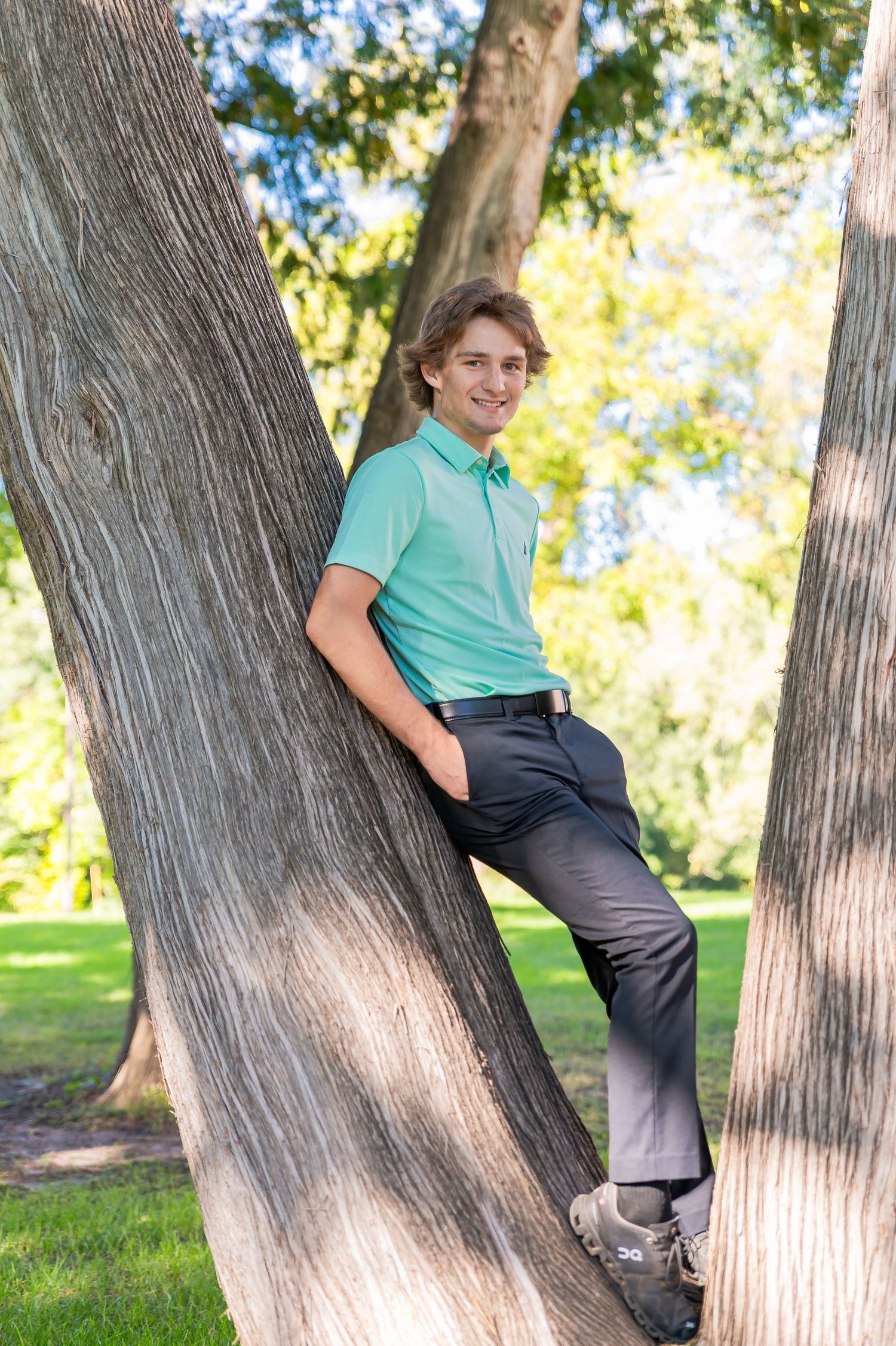 A person in a mint green shirt and dark pants sits casually on a leaning tree trunk in a sunny park setting.