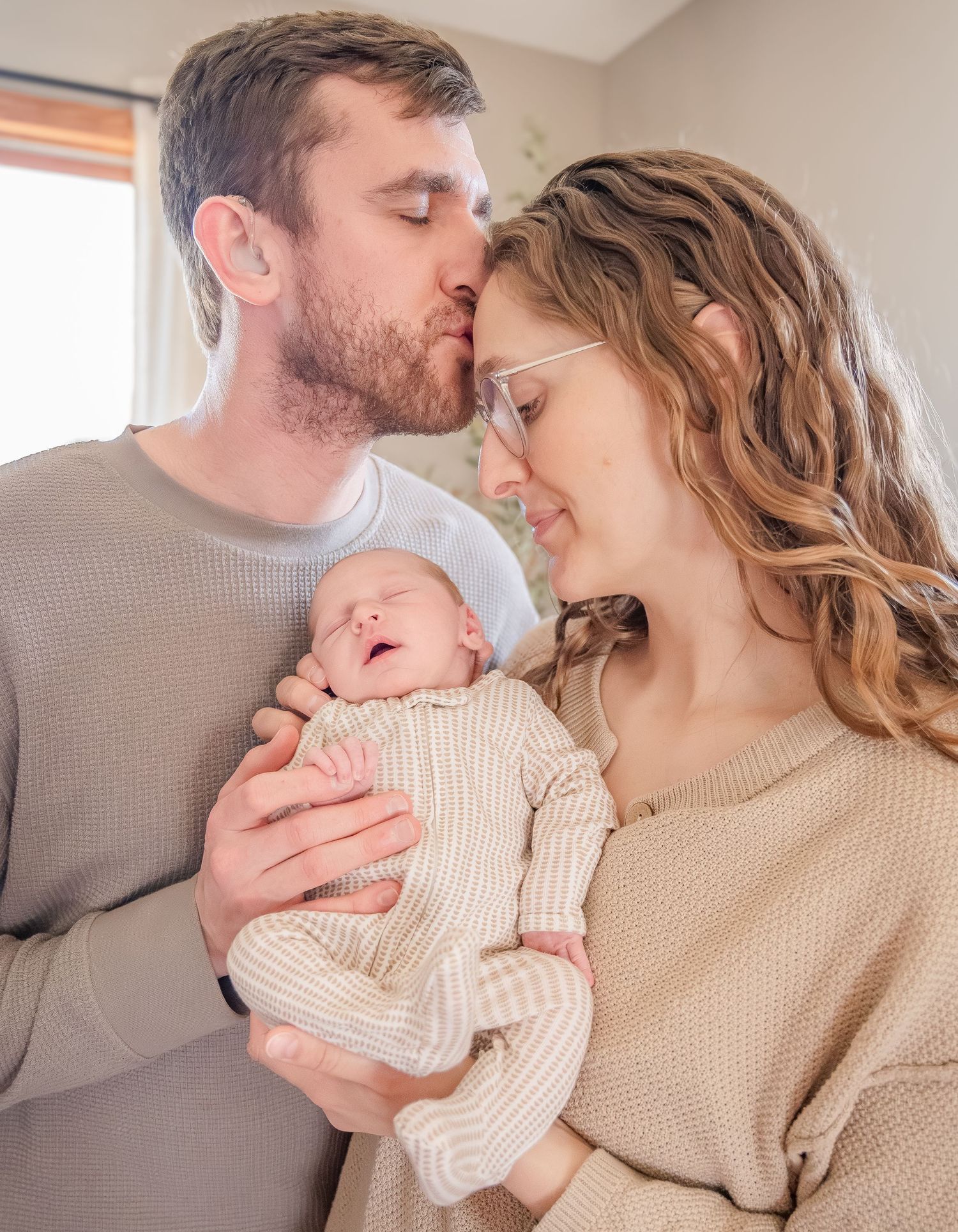 Parents share tender moments with their newborn baby while wearing neutral colored sweaters in a bright room.