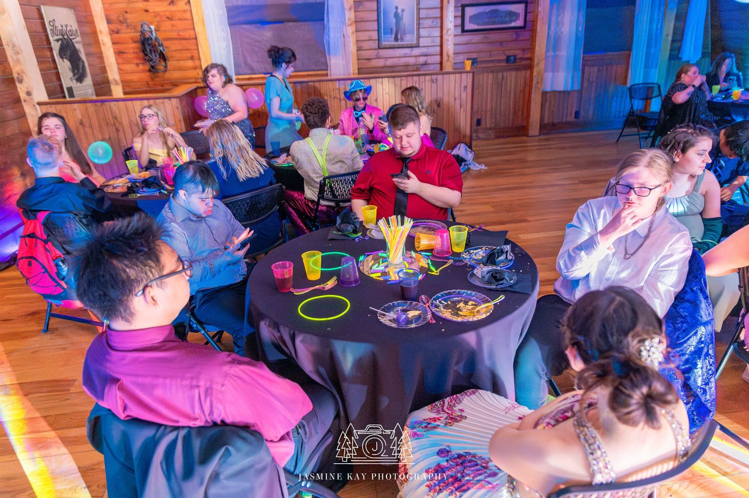 Guests seated at round tables with colorful decorations during an indoor celebration event in a wood-paneled room.