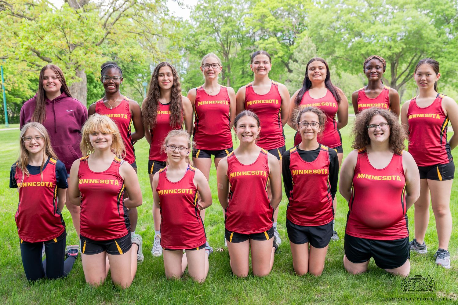 A group of athletes in red jerseys pose together outdoors on grass for a team photo.