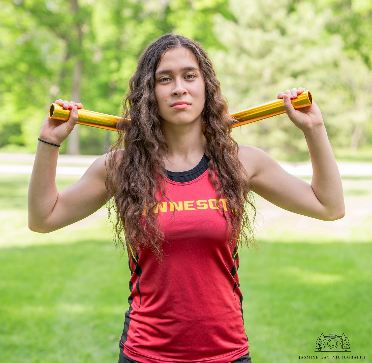 Athletic person in red tank top holding resistance bands outdoors in sunny park setting.