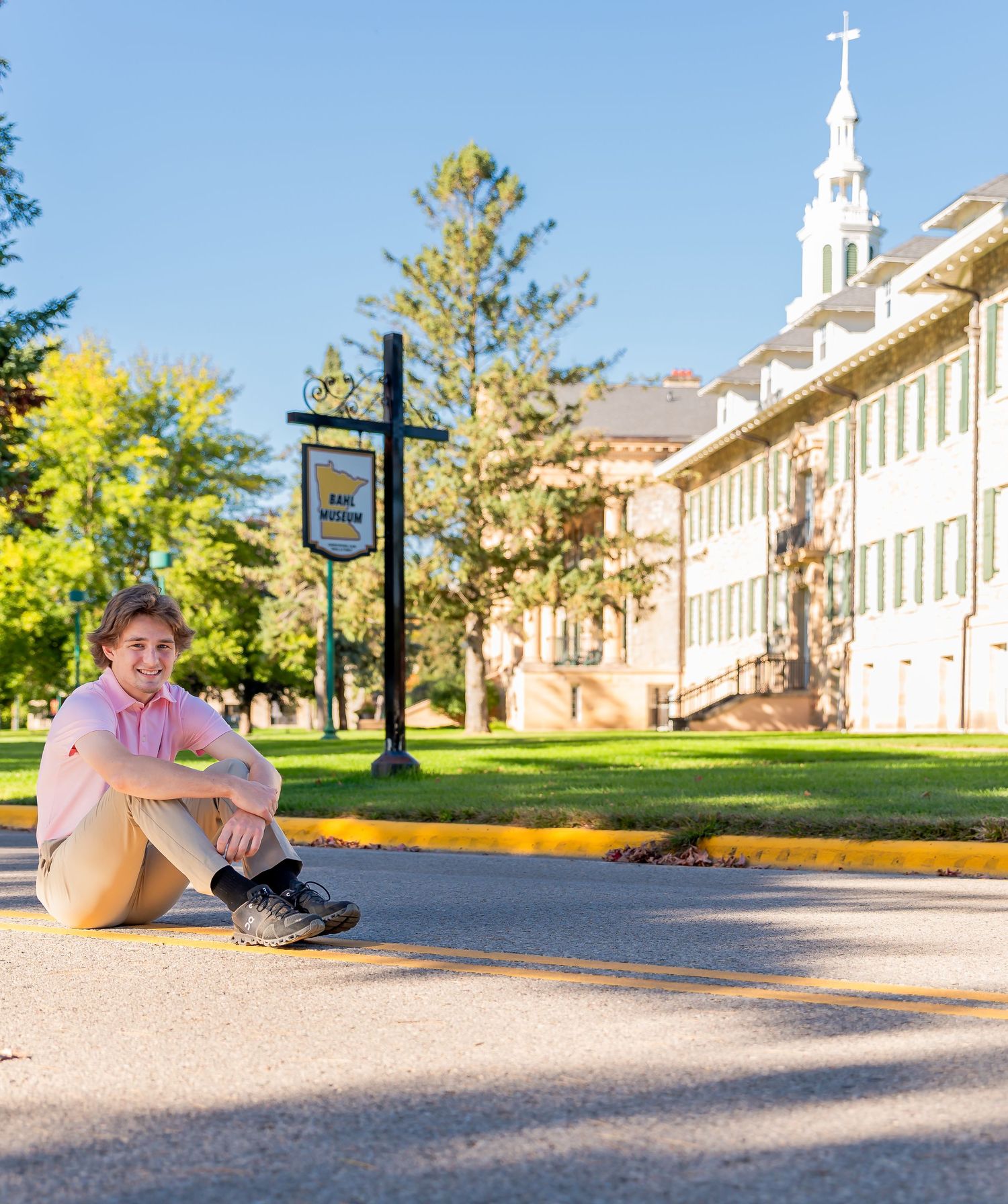 Student sits on a curb near a traditional college building with white steeple and autumn trees on campus.