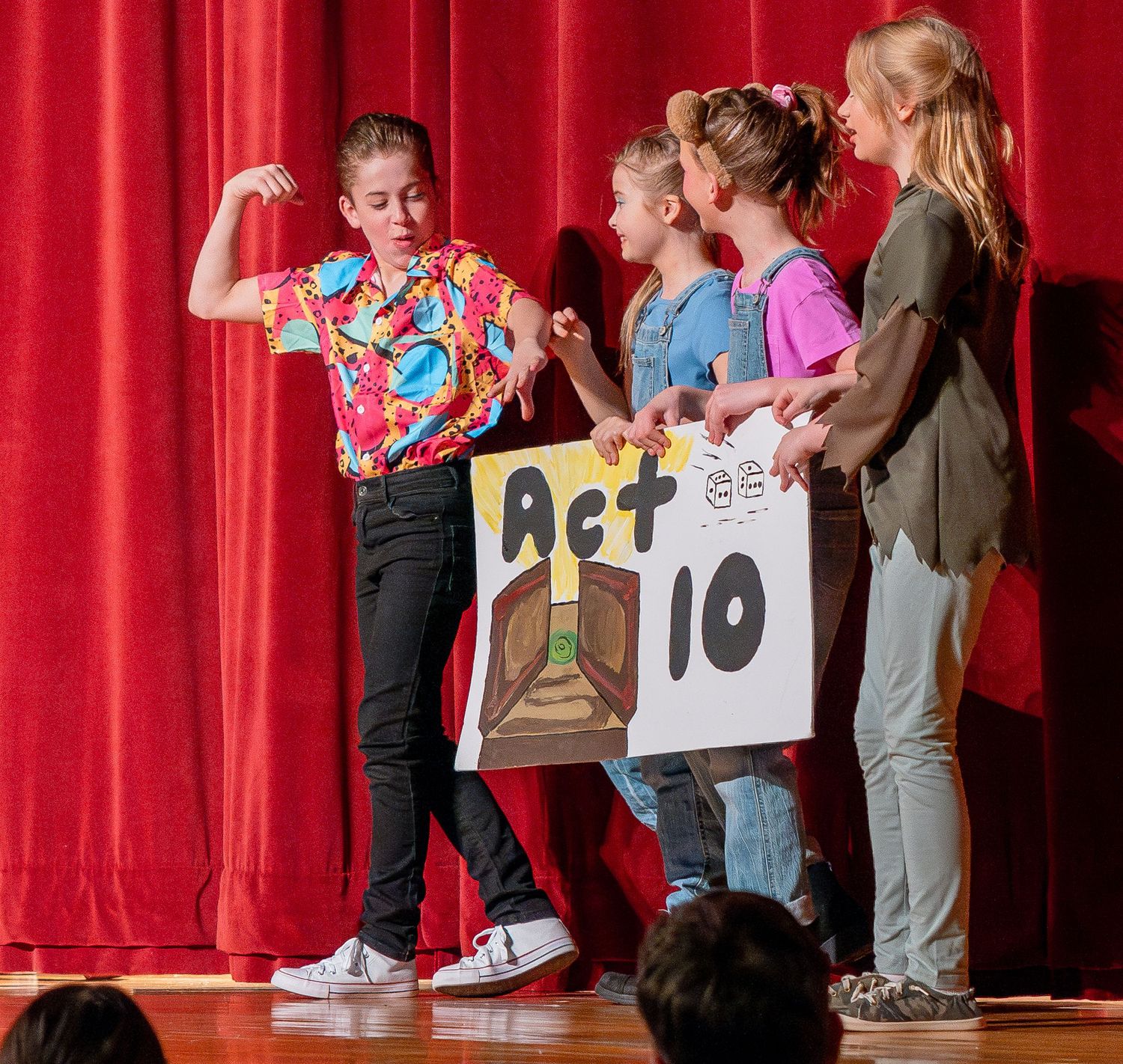 Students perform on stage in front of red curtains during a school production.