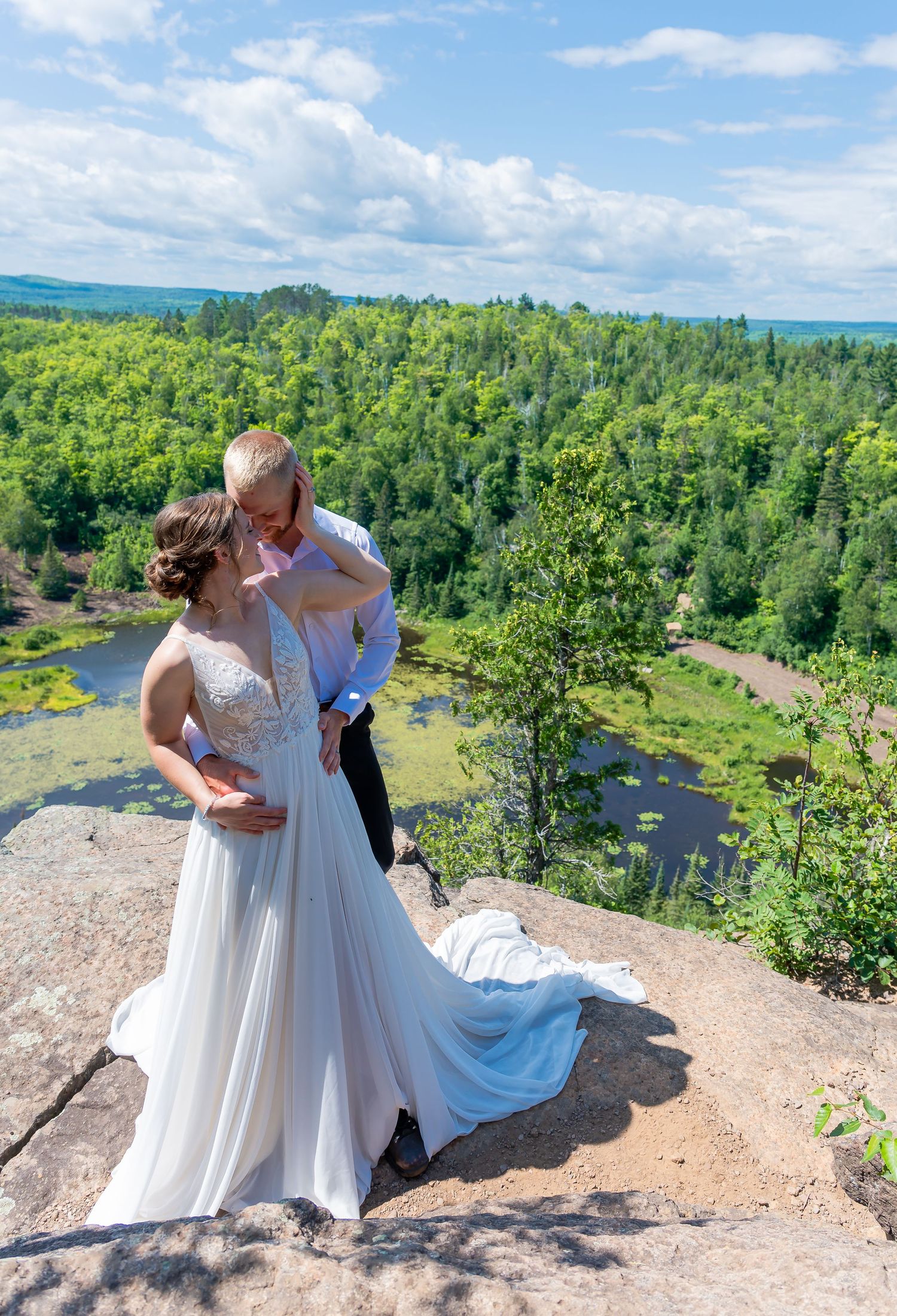 Wedding couple embracing on cliff overlook with dramatic forest valley view.