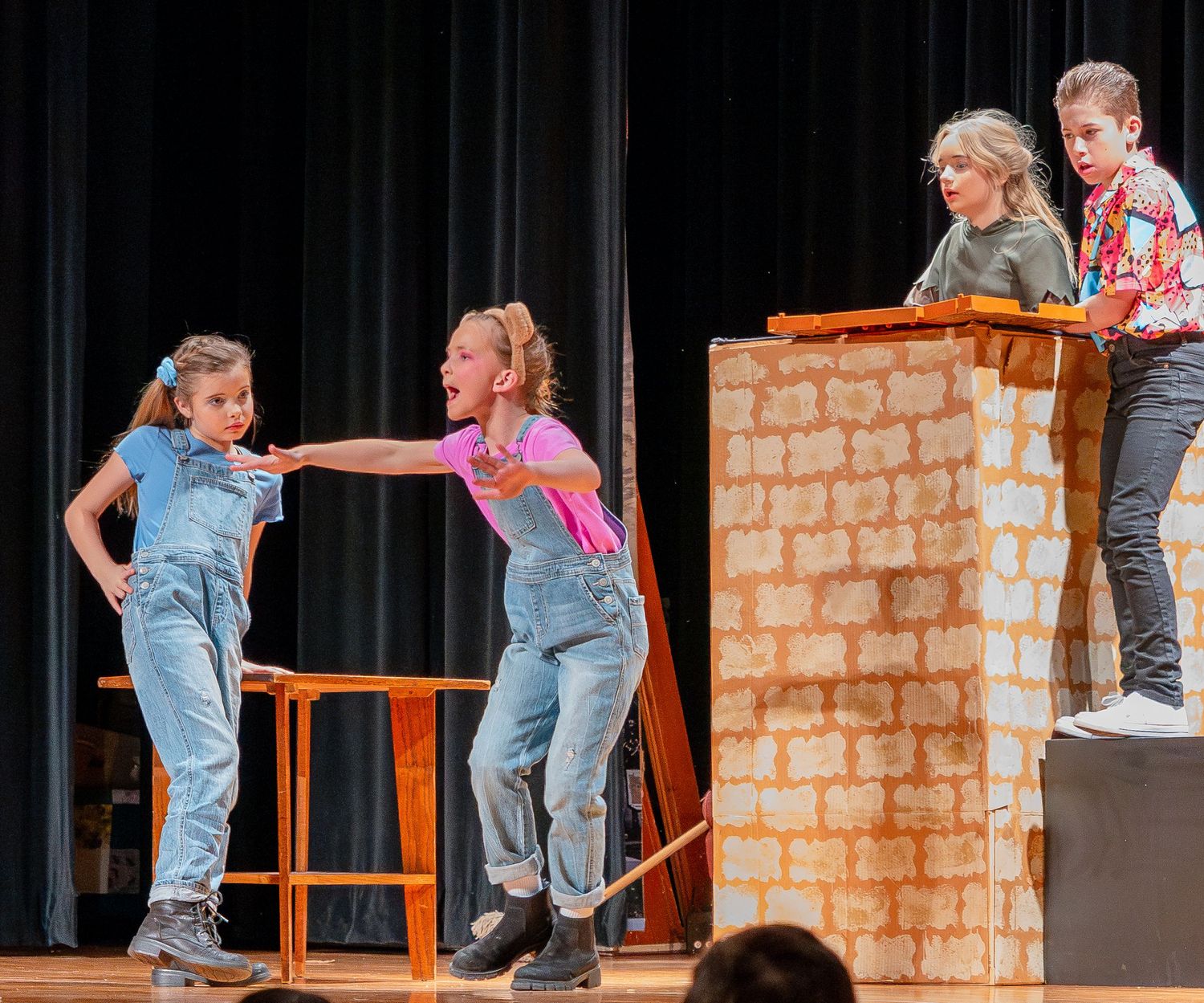 Children perform an energetic scene on stage with wooden furniture and brick wall backdrop during a theatrical production.