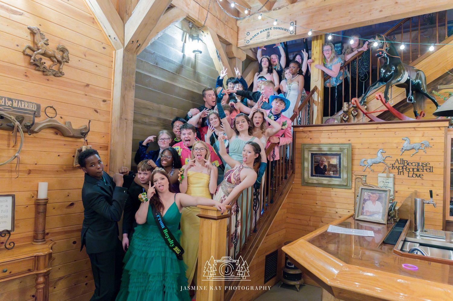 Wedding party gathers around a curved wooden staircase in a rustic log cabin venue for formal photos.