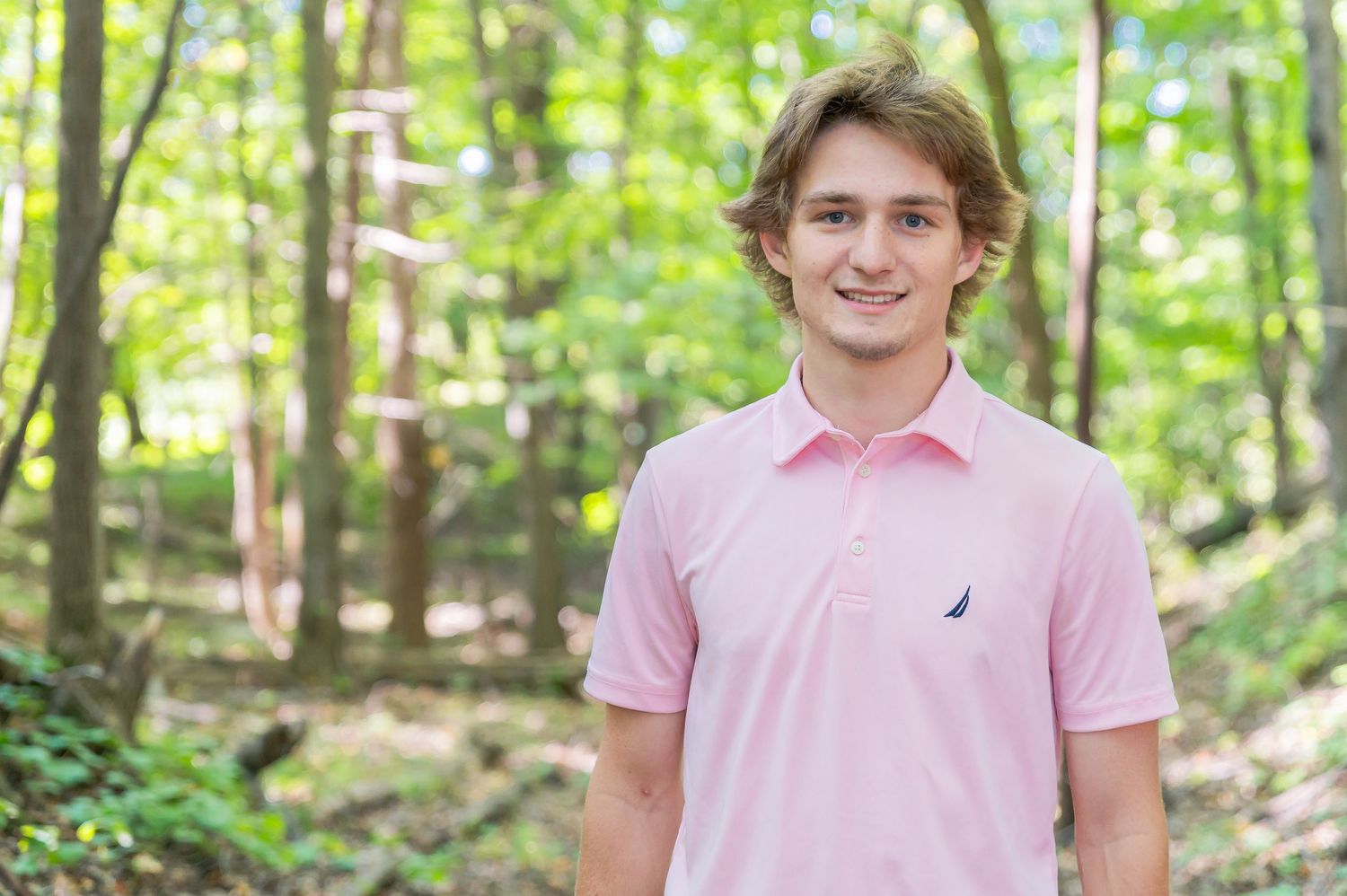 A person in a pink polo shirt with a small logo stands smiling in a sunlit wooded forest setting.