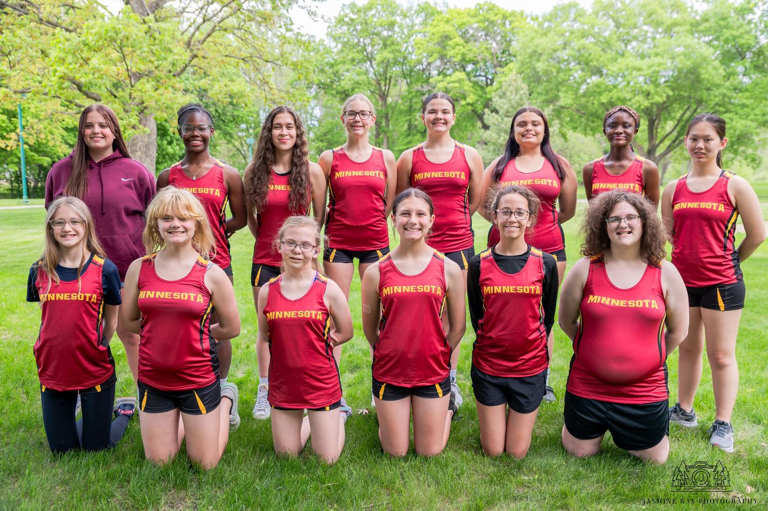 A sports team wearing red jerseys poses together on grass for a group photo in a park setting.