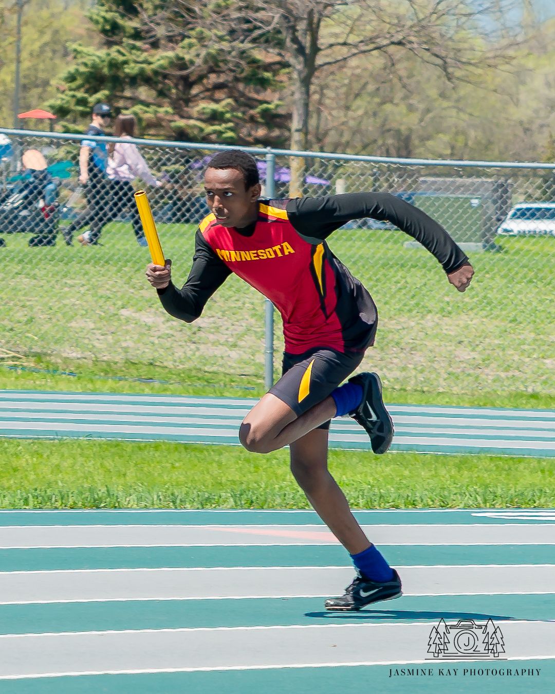 Runner in red and black uniform sprinting with yellow baton on track field.