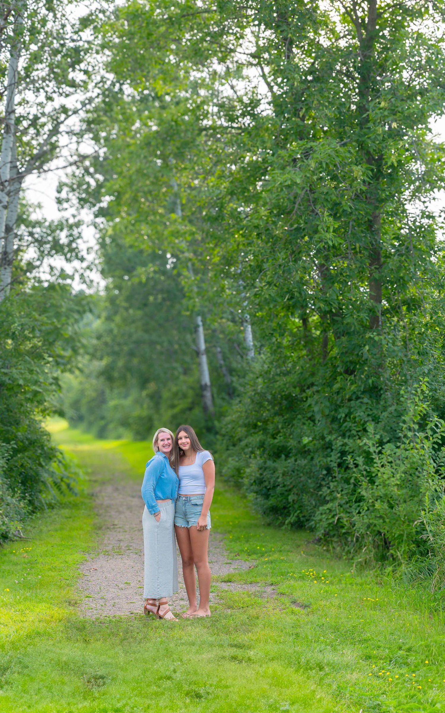 Two people stand on a grassy path surrounded by lush green trees and foliage on a summer day.