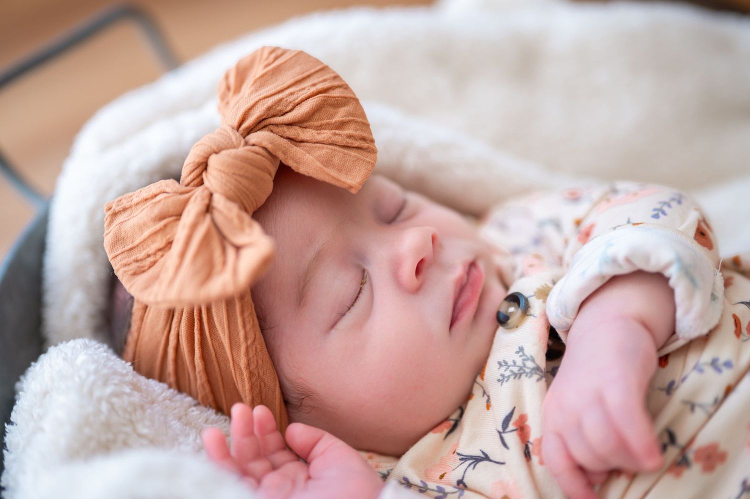 Sleeping baby wearing floral outfit and peach-colored bow headband rests peacefully on soft white blanket.