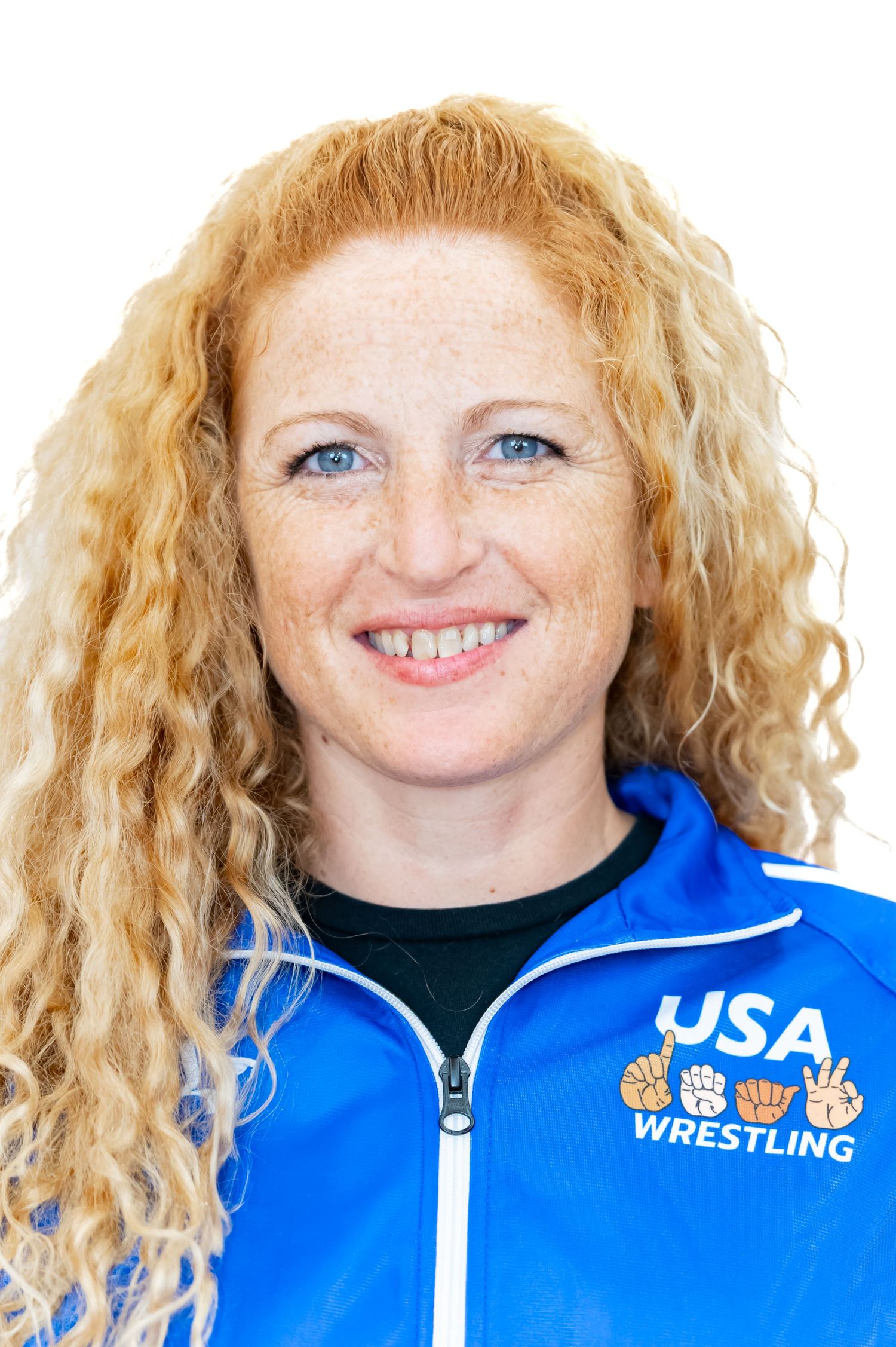 A person in a blue USA Wrestling jacket smiles at the camera with curly blonde hair.