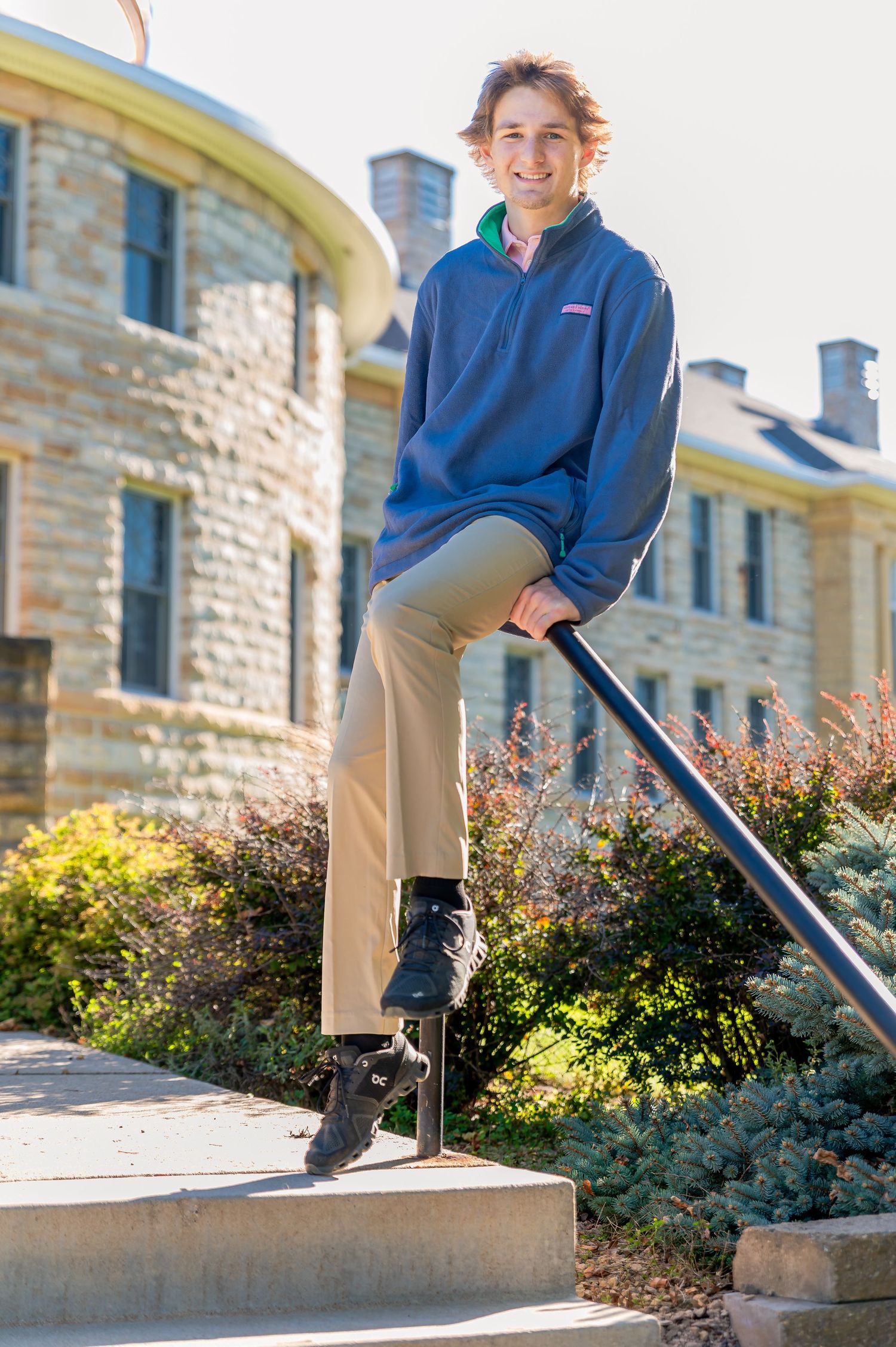 Person in blue fleece and khaki pants performs a skateboarding trick on concrete steps near a stone building.