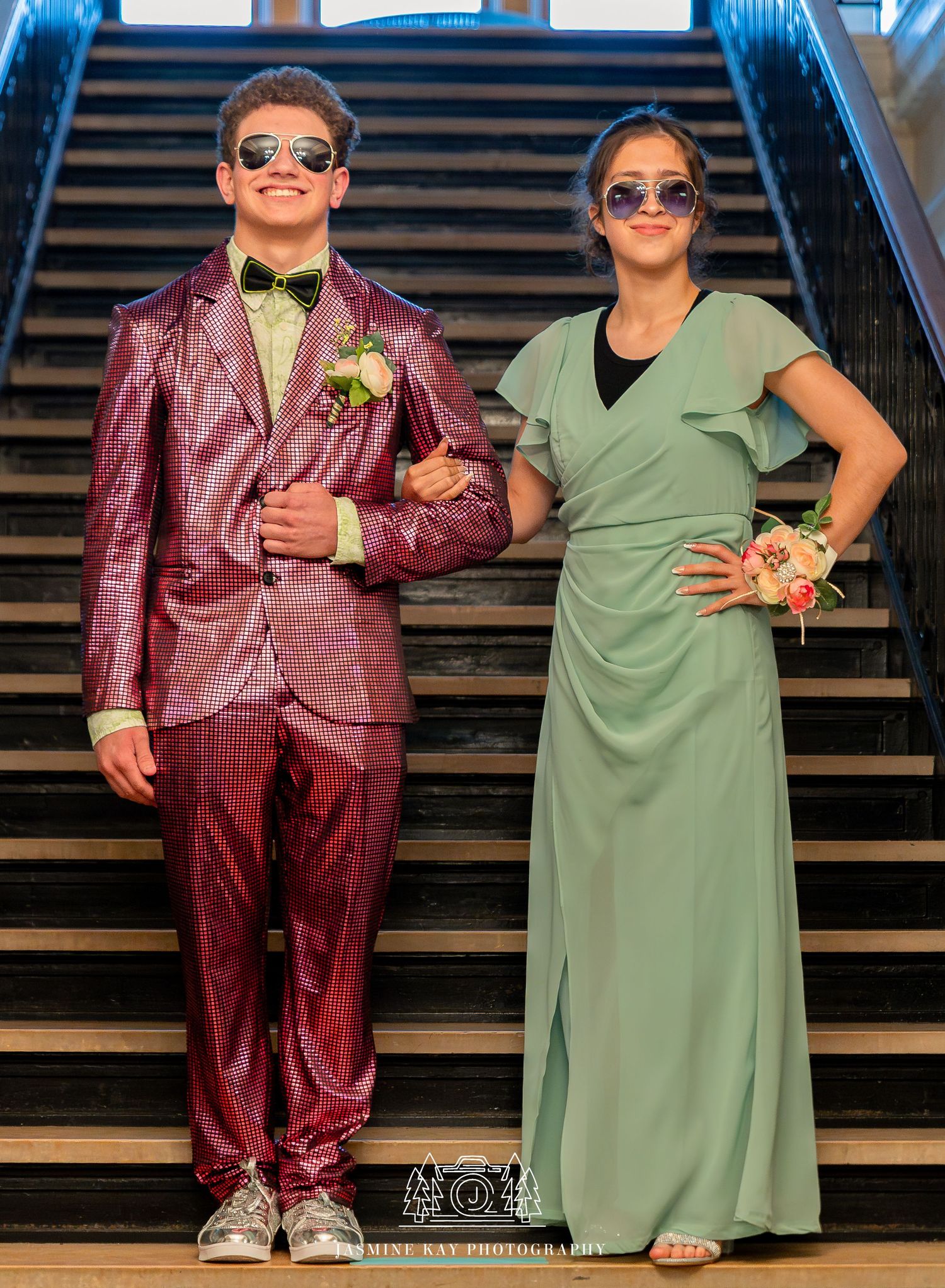 Two people in contrasting outfits, one in burgundy suit and one in sage dress, pose together on stairs.