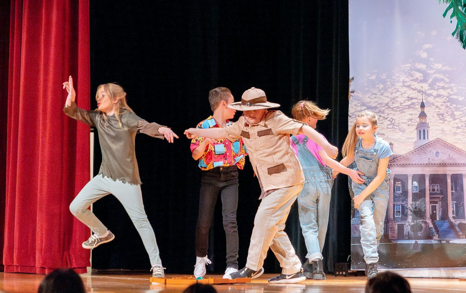 Dance ensemble performs a western-themed choreography on stage with red curtains and scenic backdrop.