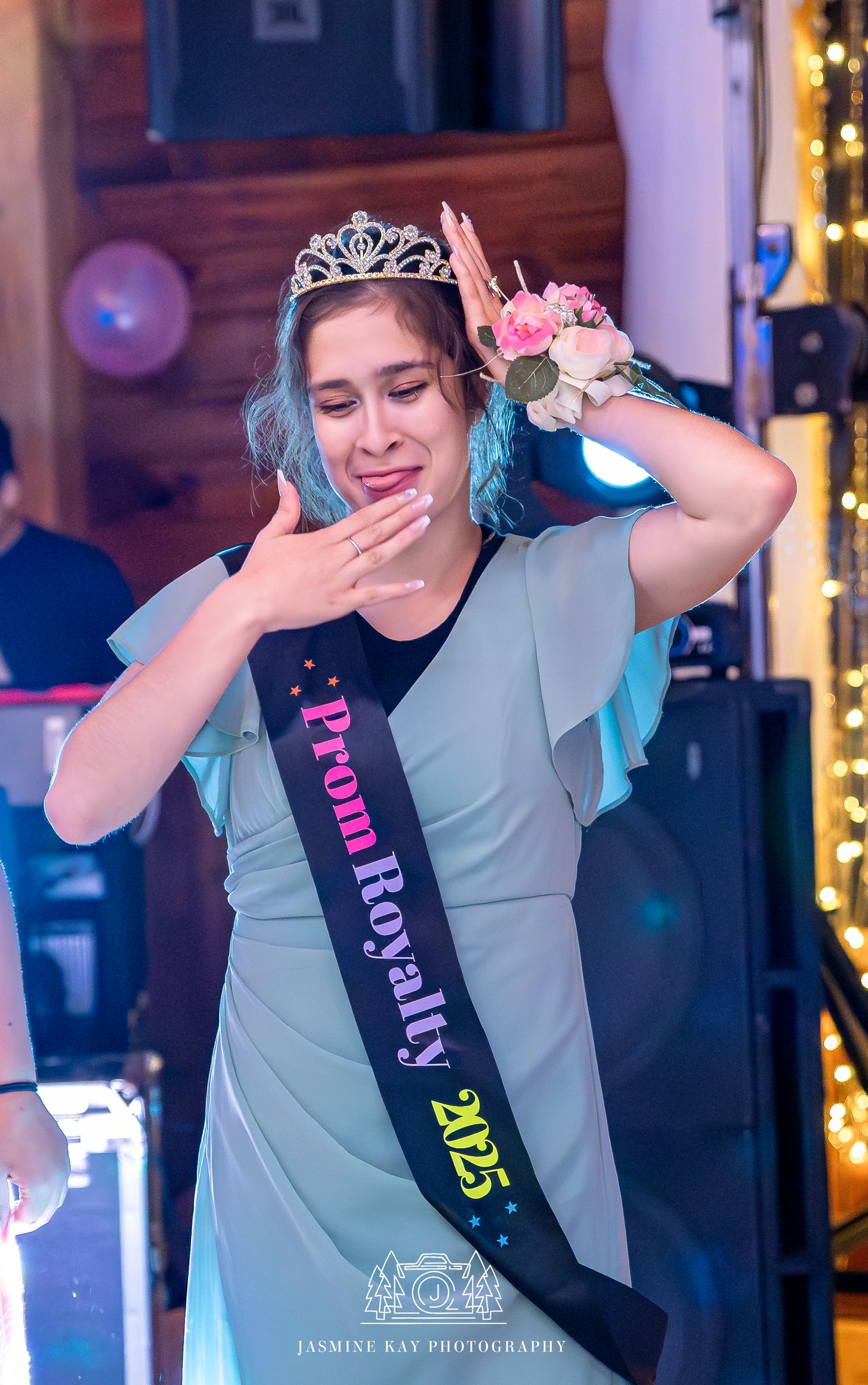 A person wearing a tiara and prom royalty sash poses emotionally at a formal event with blue hair and floral wrist corsage.