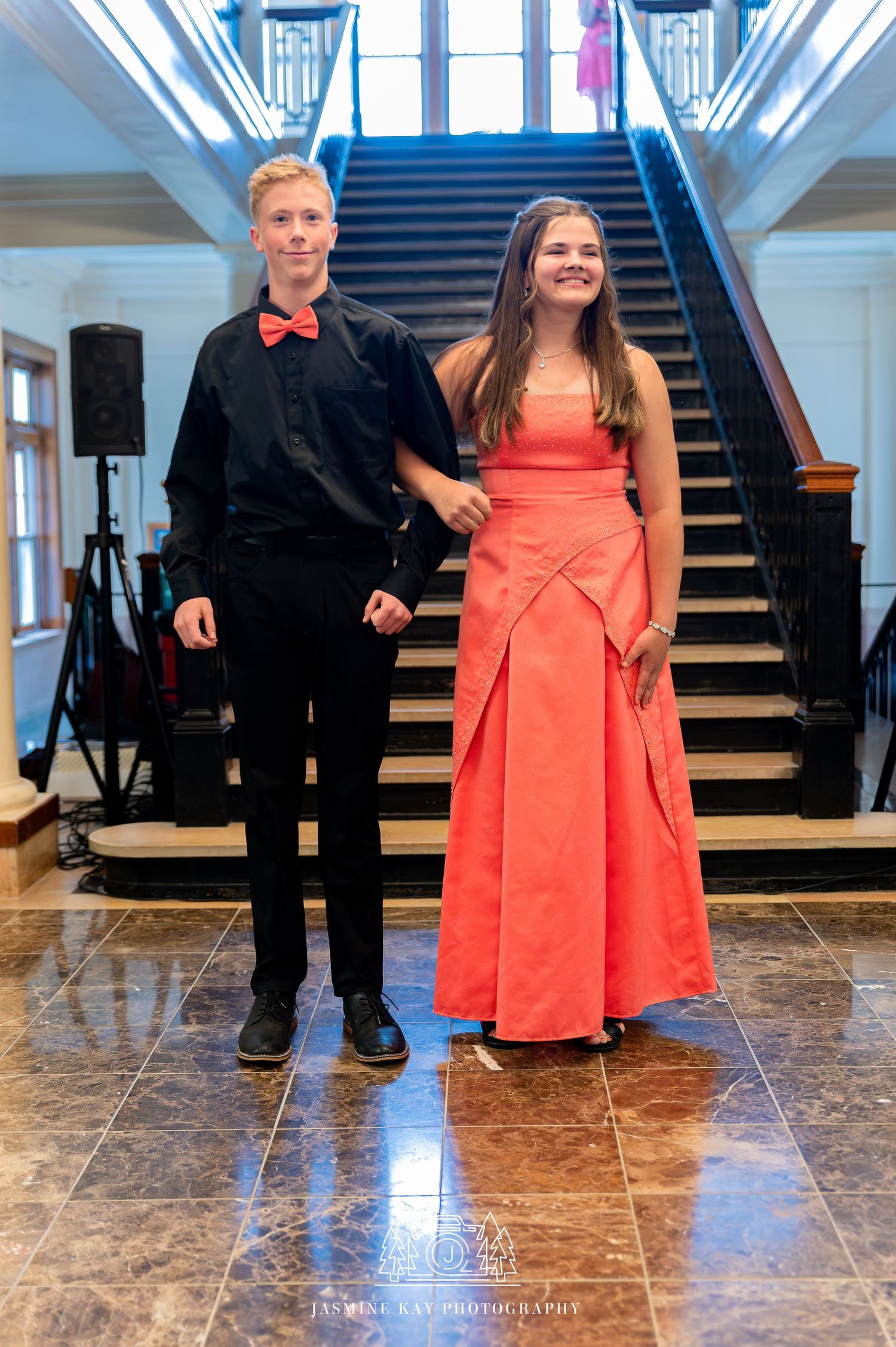 Couple in formal attire with black suit and coral dress posing on marble floor near grand staircase.