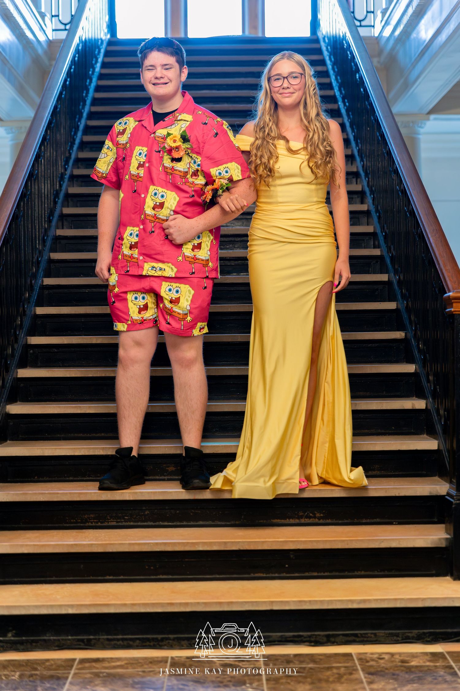A couple poses on stairs, wearing coordinating yellow dress and SpongeBob print outfit in an indoor setting.