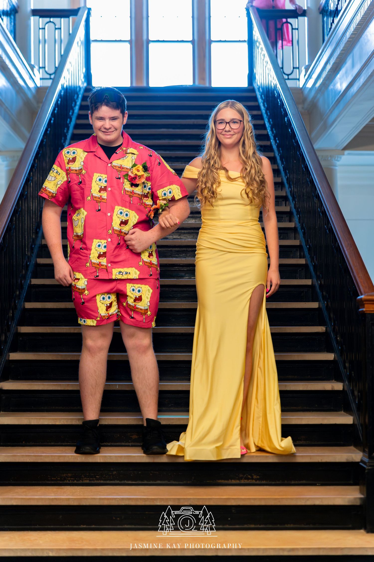 Two people pose on stairs, one wearing a bright red patterned romper and another in a flowing yellow formal gown.