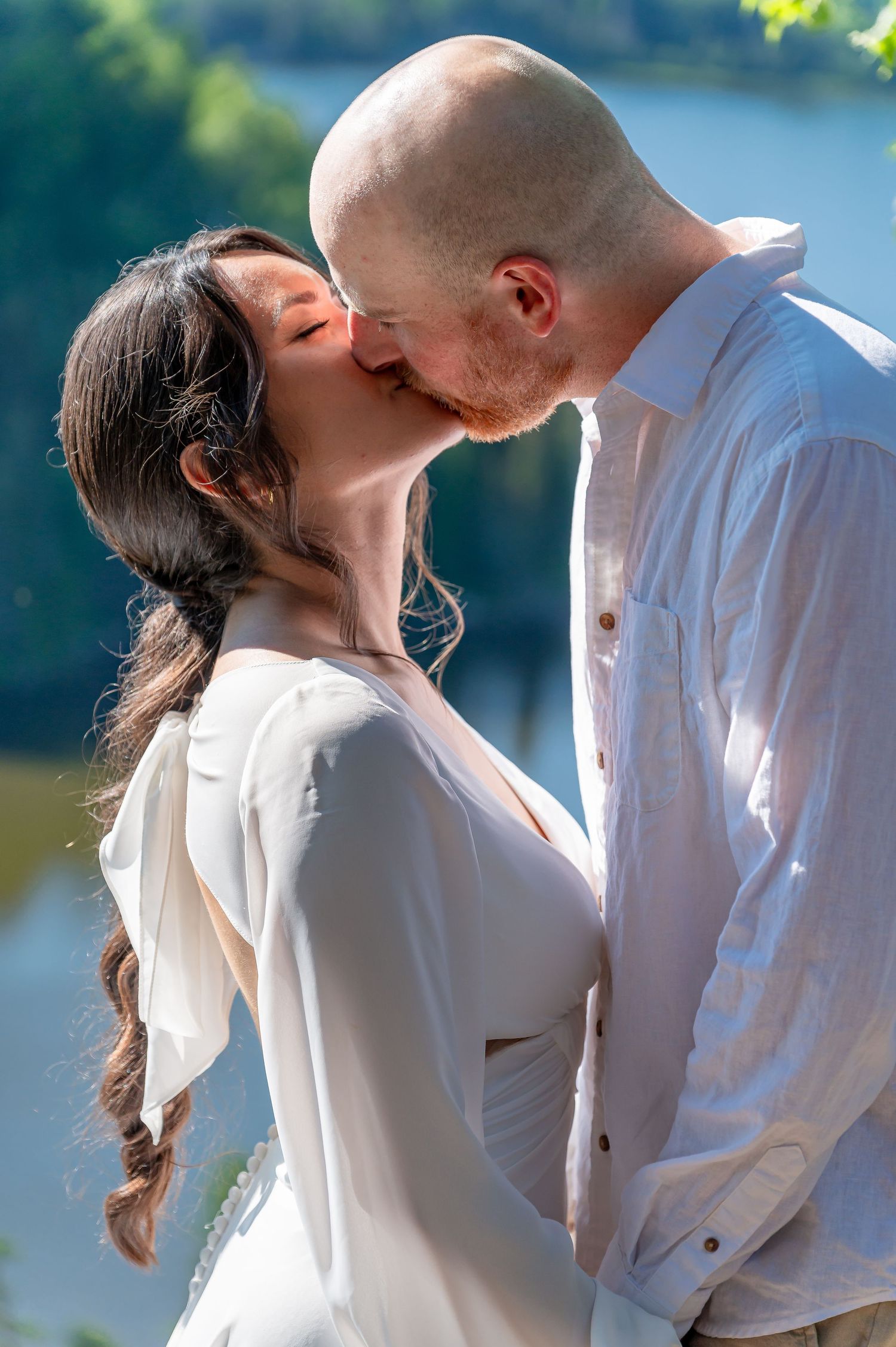 Intimate couple portrait with white flowing attire against scenic lake and forest background.
