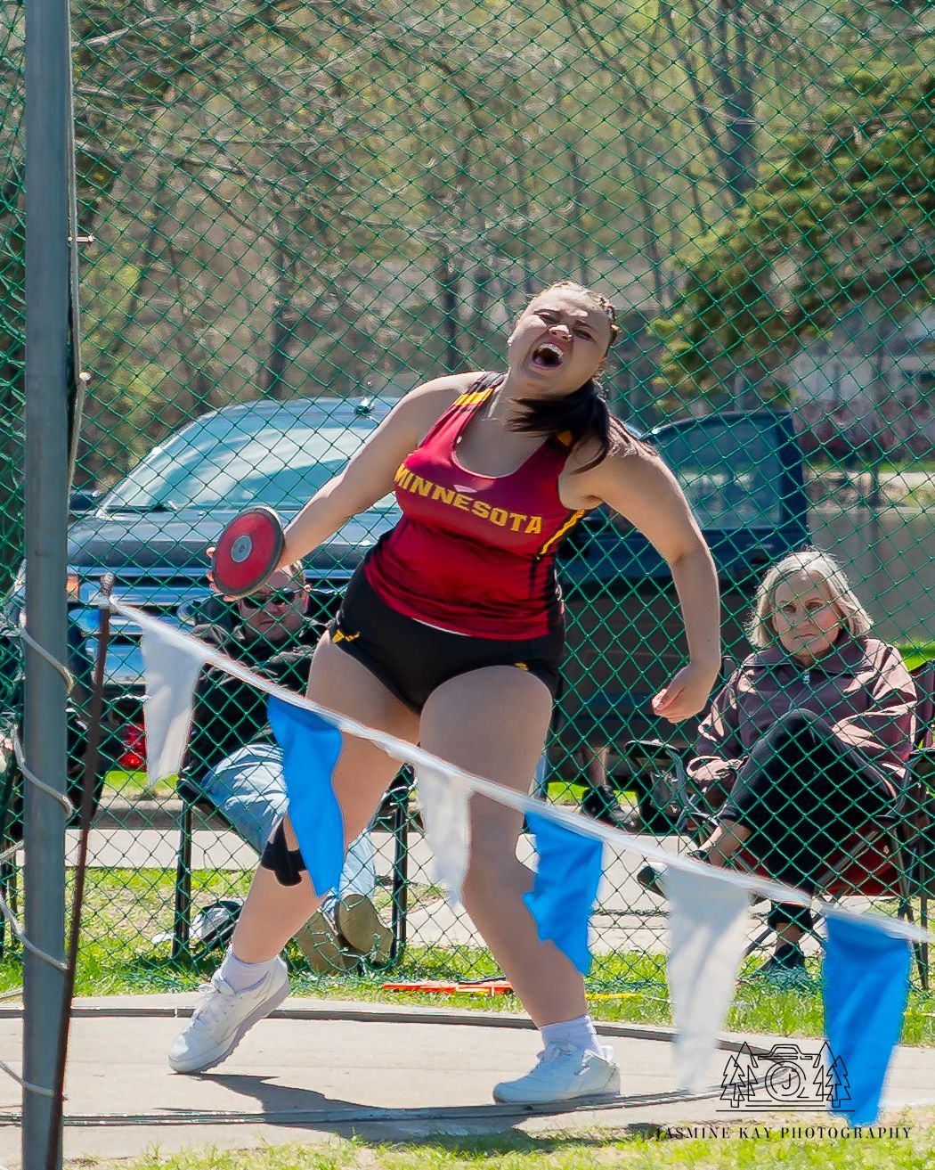 Athletes in red uniforms stretching and warming up on an outdoor track field with chain link fence in background.