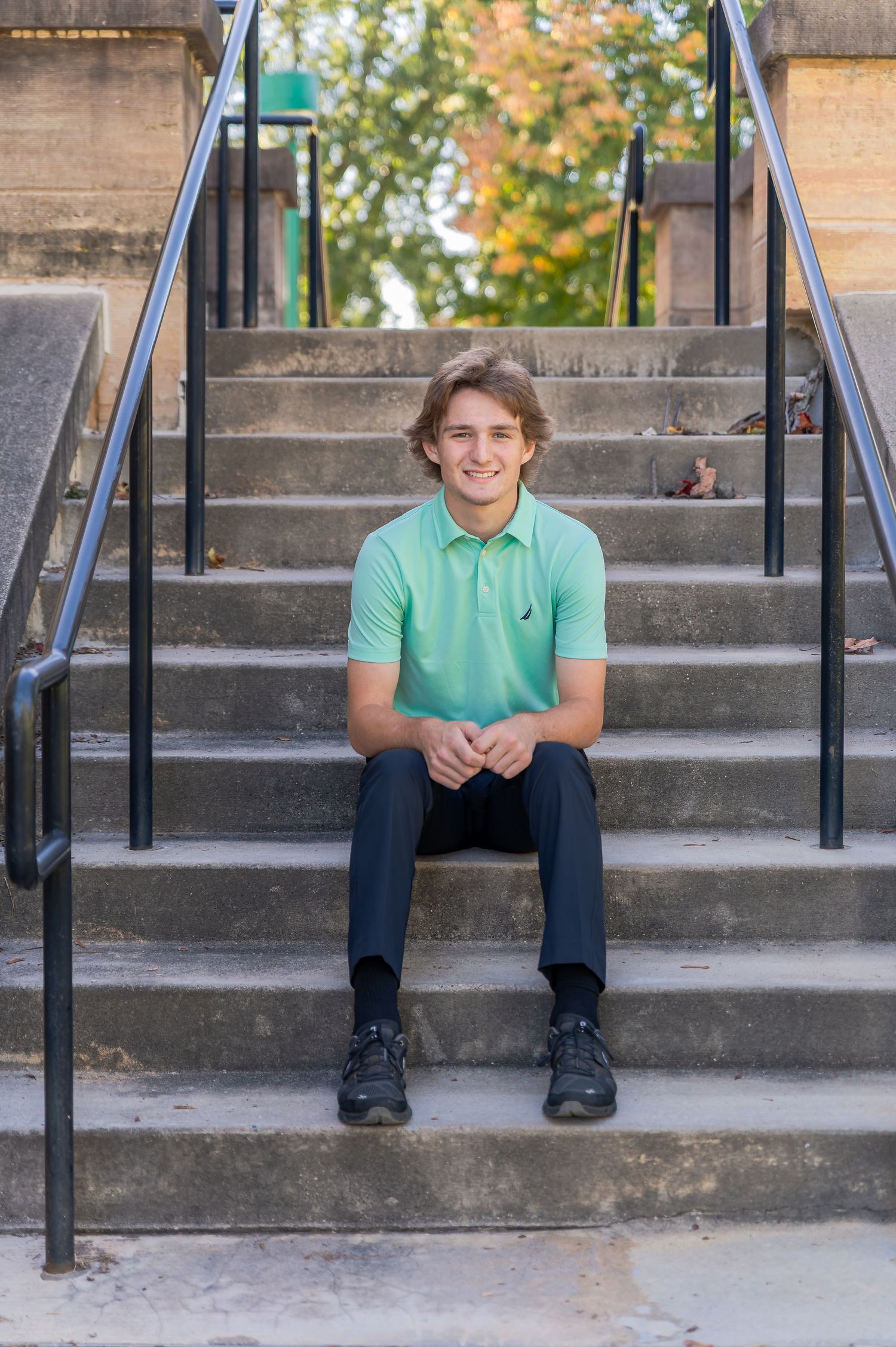Young student in mint green polo shirt and dark pants sits on concrete steps outdoors in autumn.