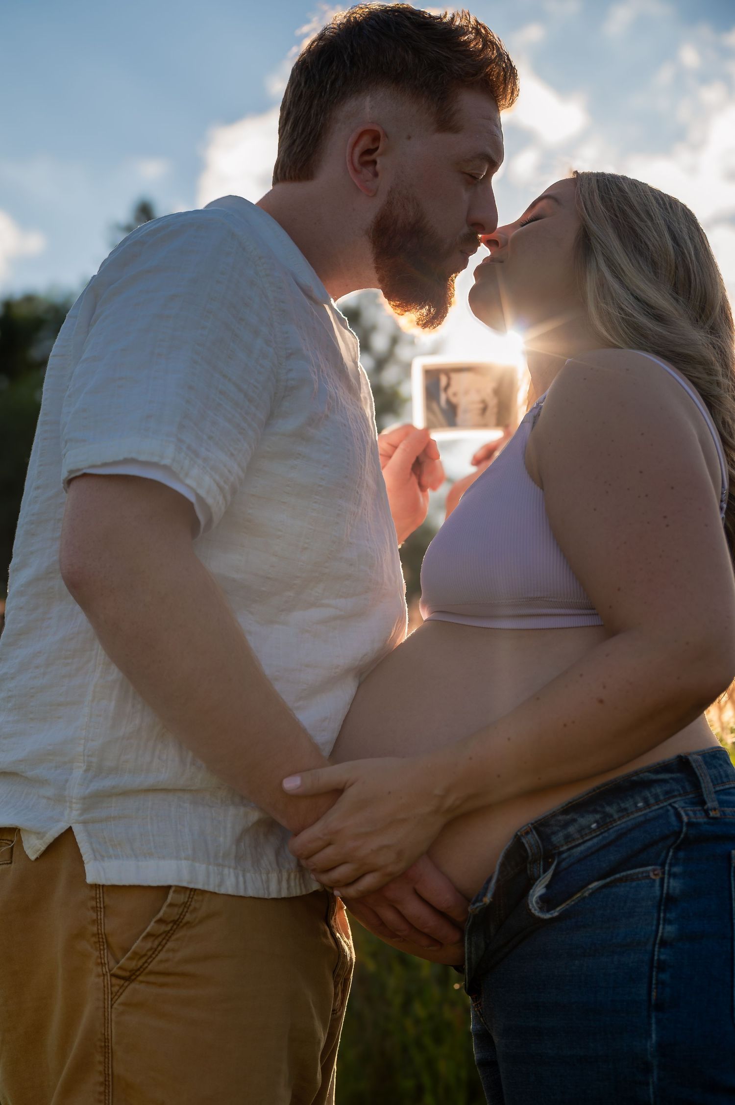 A romantic couple embraces in a golden sunset field wearing casual clothing as the sun glows behind them.