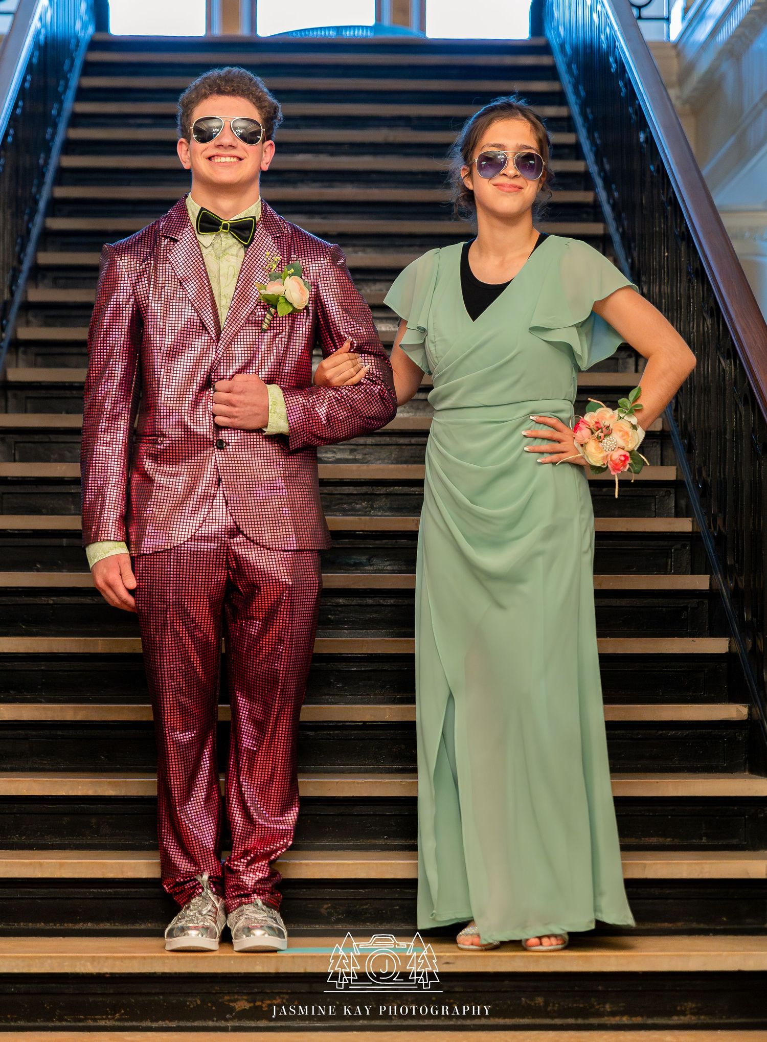 A couple in contrasting burgundy suit and sage green dress pose playfully on wooden stairs wearing sunglasses.