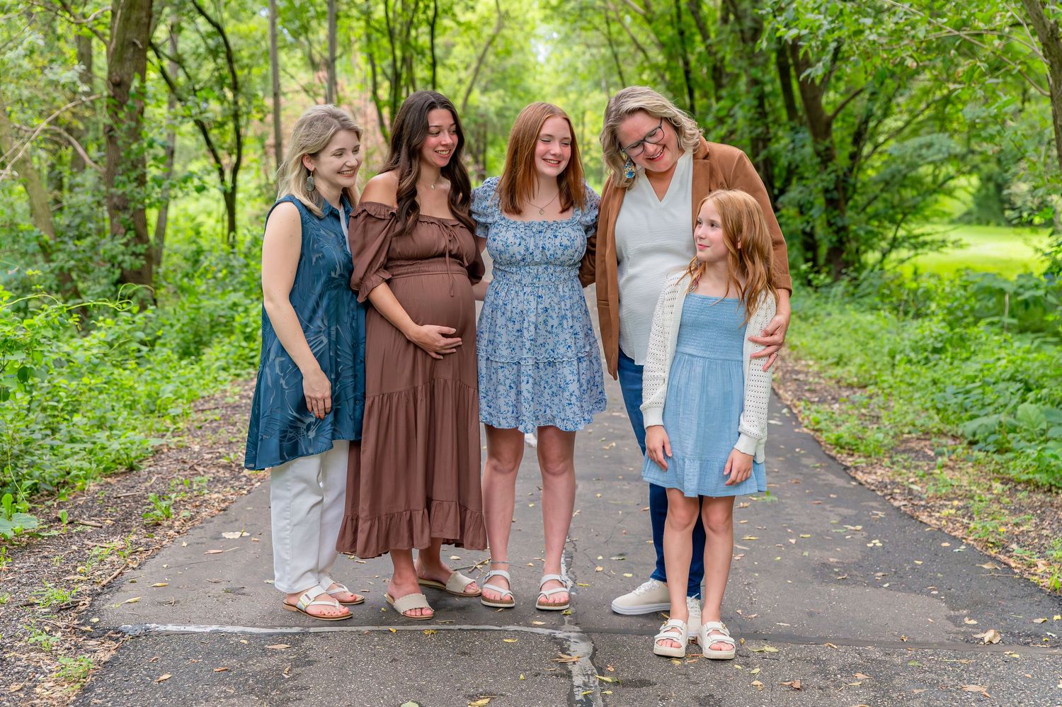A group of friends in coordinating brown and blue dresses pose together on a tree-lined path.