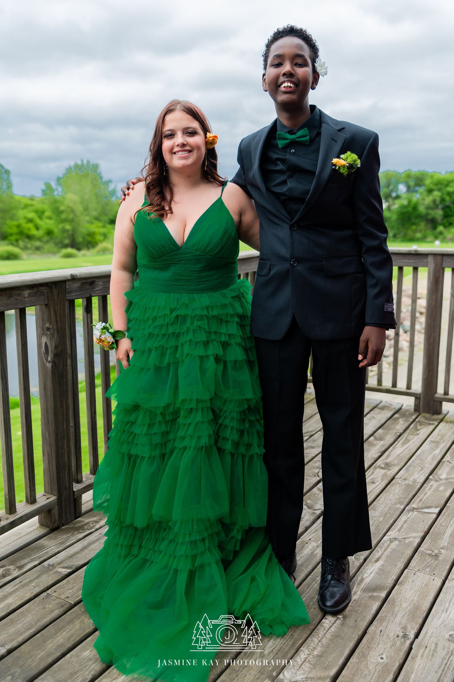 A couple in formal attire, with a bright emerald green tiered dress and black suit, pose together on a wooden deck outdoors.