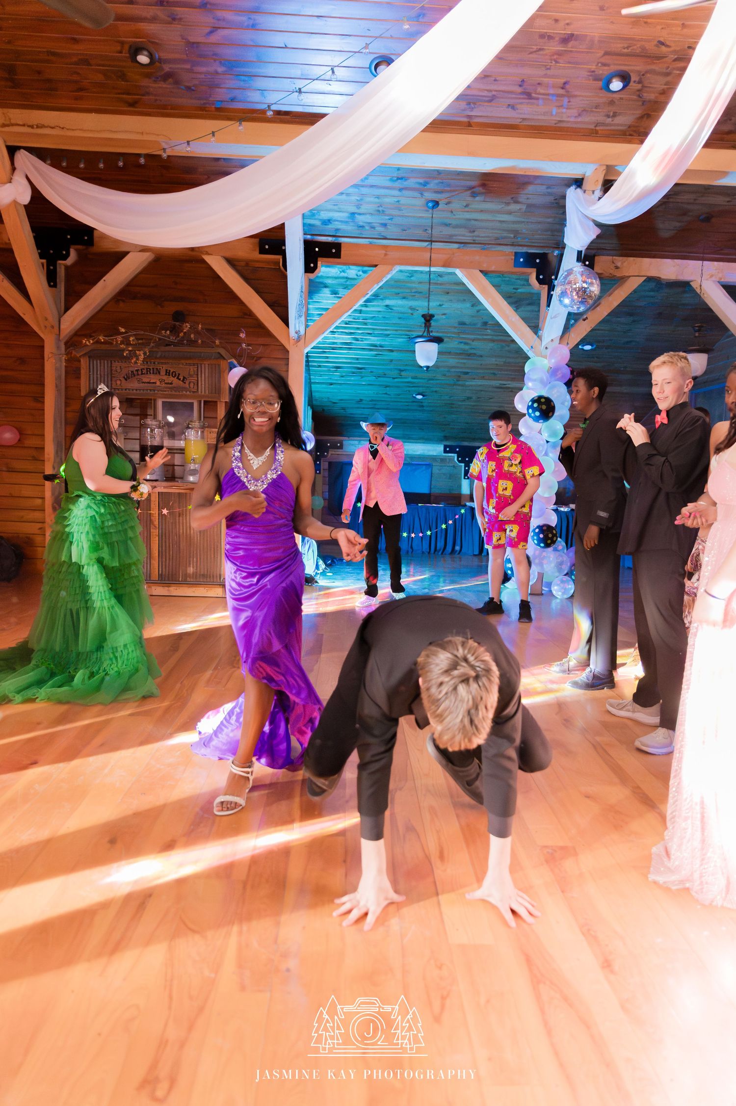 Wedding guests dance together on a polished wooden floor in a rustic barn venue decorated with white fabric draping.