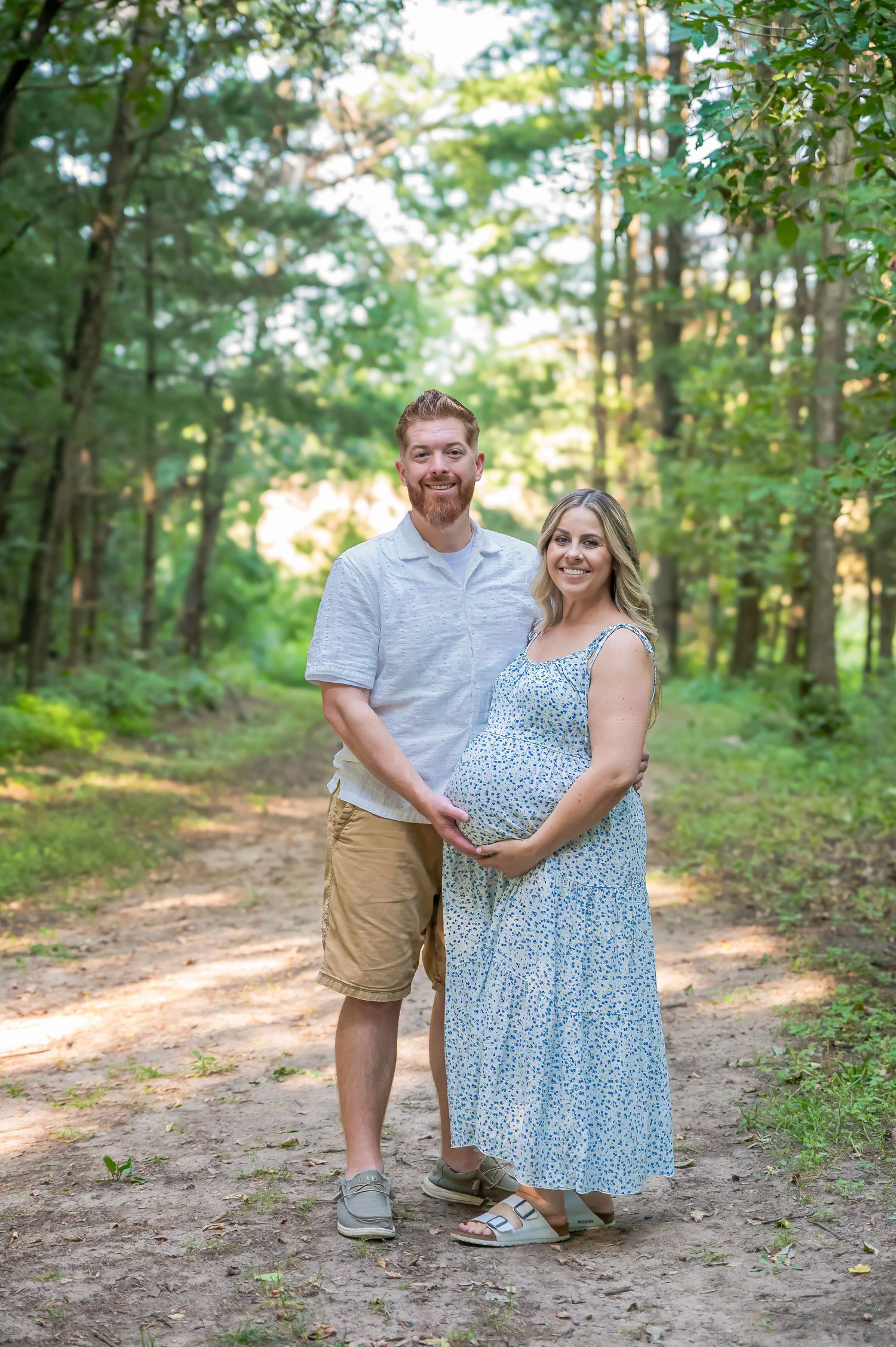 A couple poses together on a wooded dirt path during a summer photo session in a forest setting.