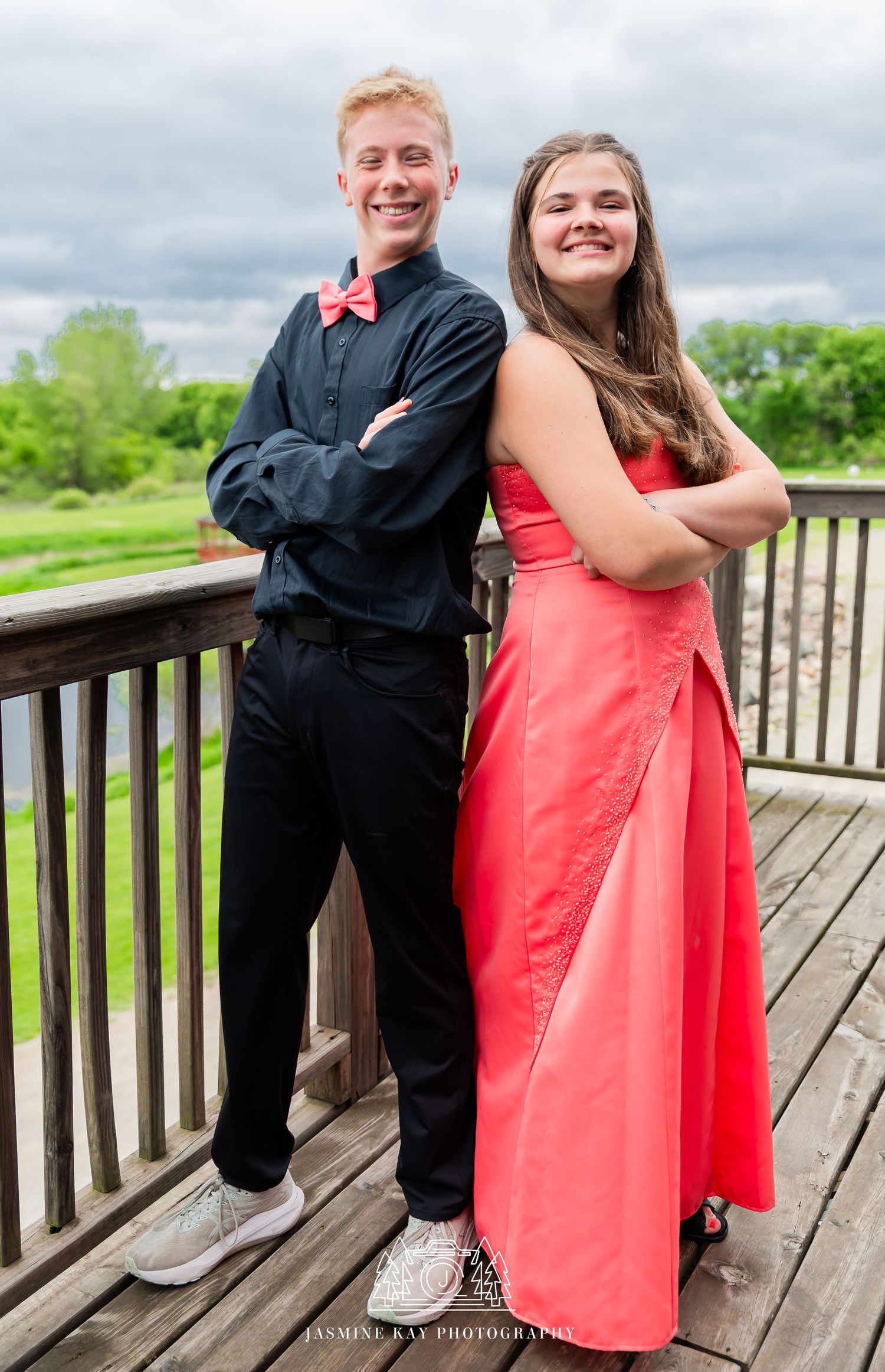 A couple in formal attire poses together on a wooden deck, with her in coral and him in black.