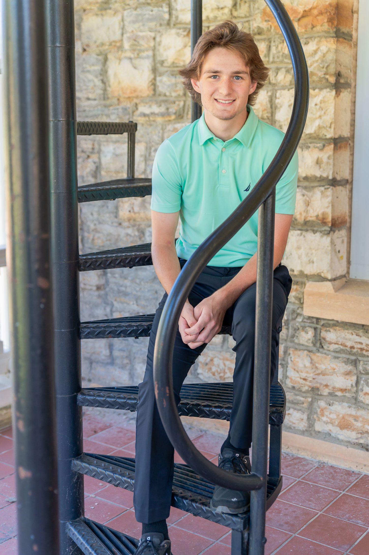 Person in mint green polo and dark pants sits casually on a stone porch with wrought iron railings.