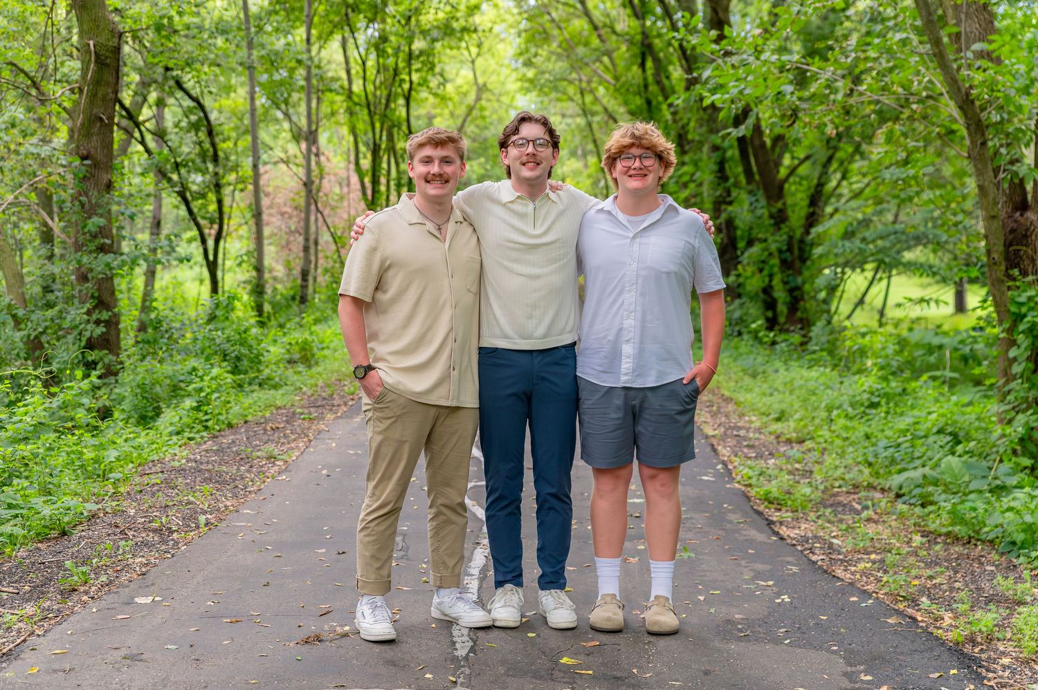 Three people stand together smiling on a tree-lined path surrounded by lush green foliage on a sunny day.