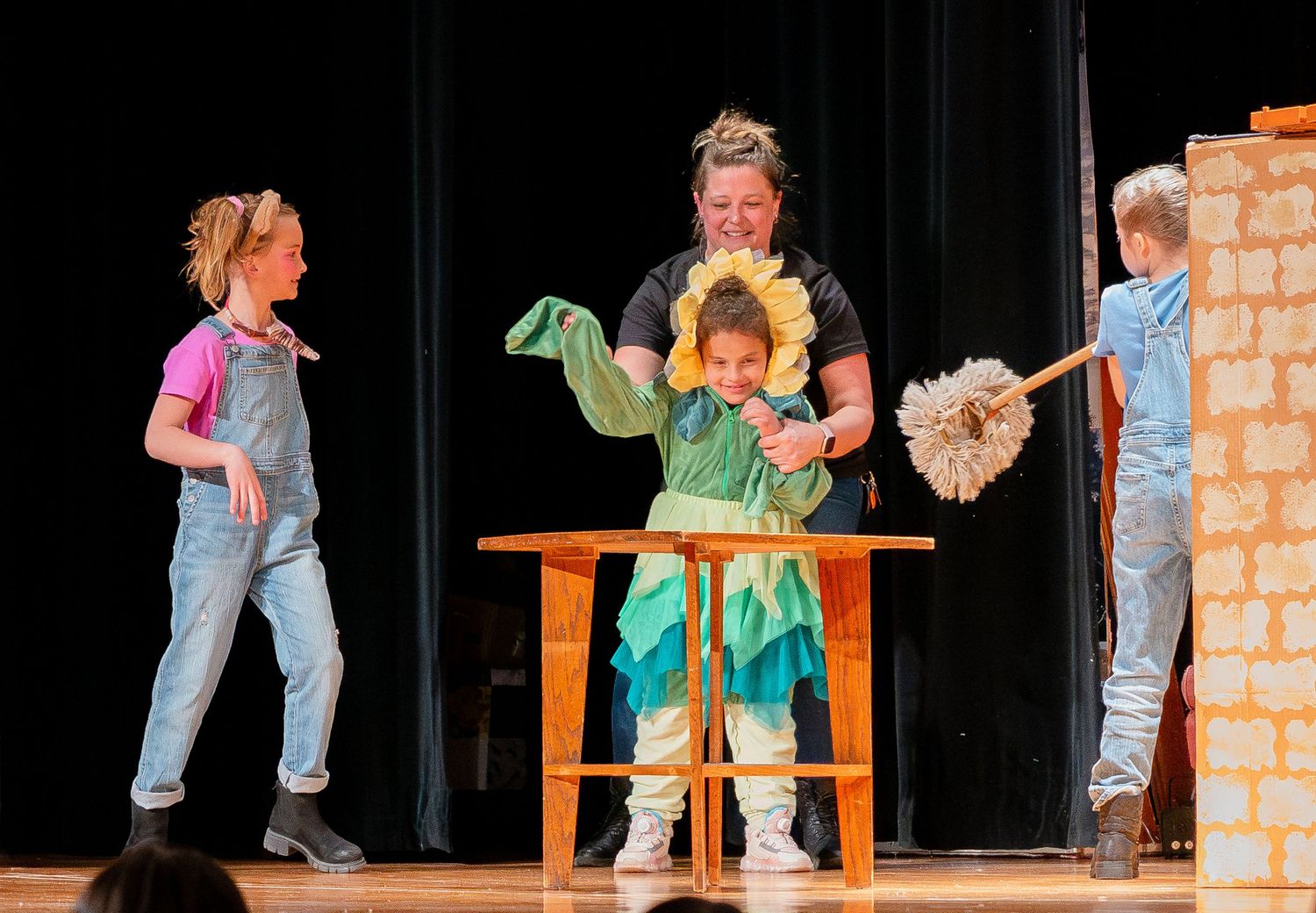 Performers in casual clothing act out scenes on a black stage with wooden furniture.