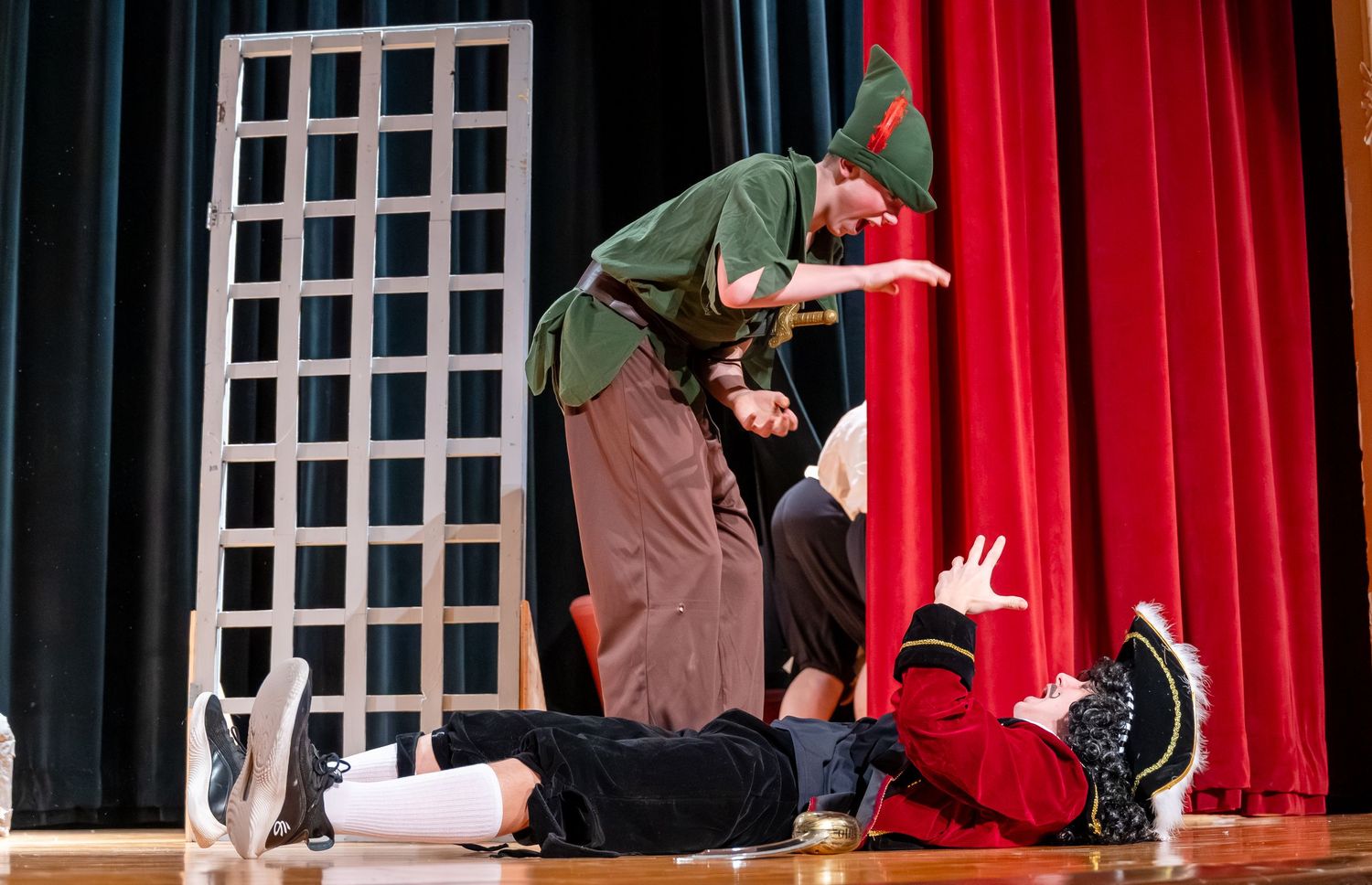 Theater performers in dark costumes act on stage with red curtains and window-like set pieces in the background.