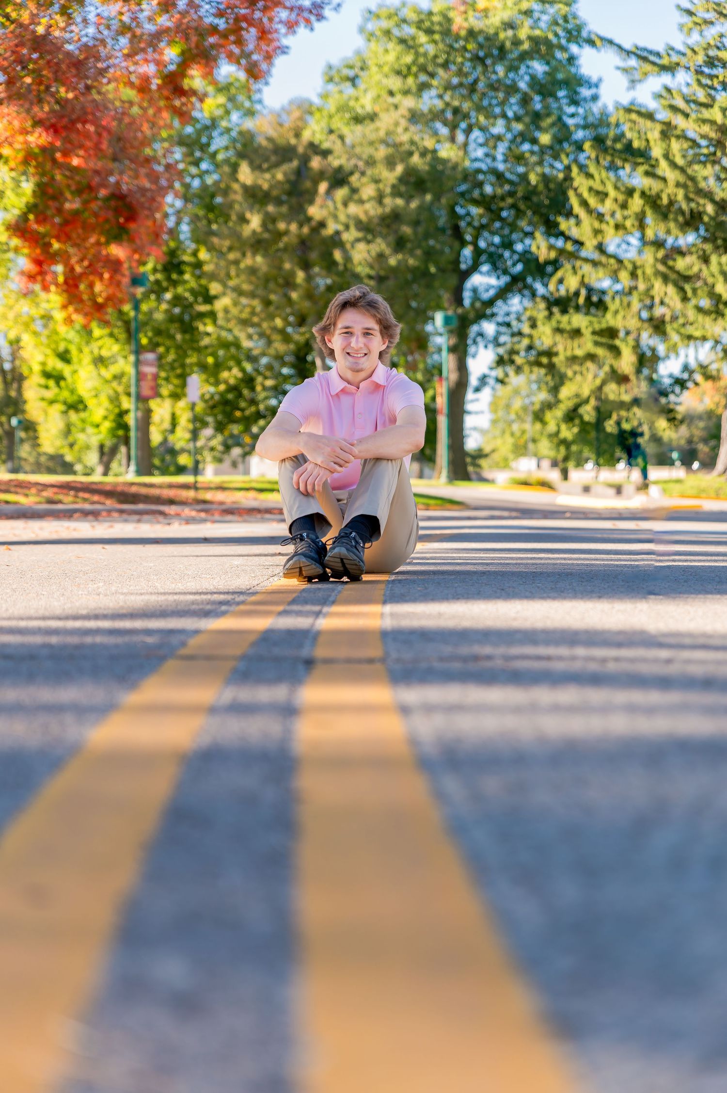 Person sitting on asphalt road with autumn trees and yellow lines casting shadows in late afternoon sunlight.