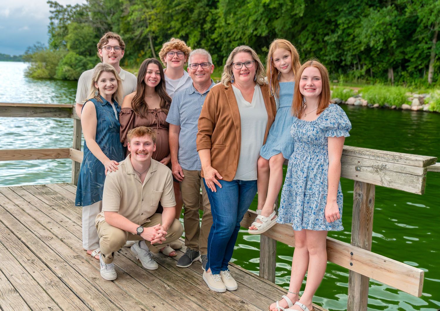 A group sits together on a wooden dock by a lake surrounded by lush green trees on a sunny day.
