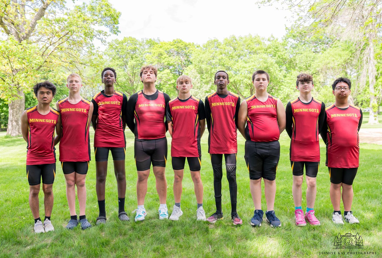 Track team members in red uniforms stand in a row outdoors against a green natural background.