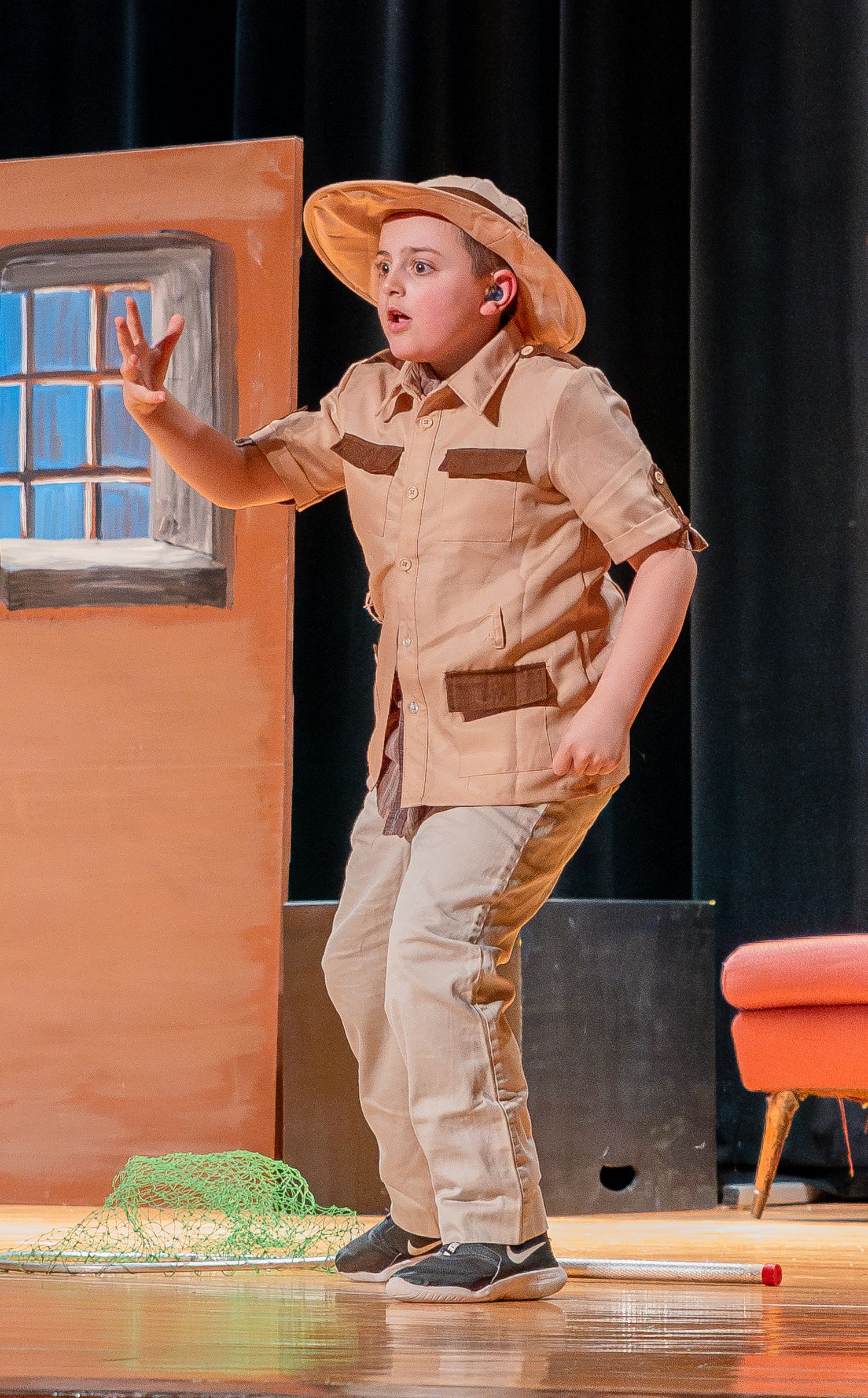 Actor in safari outfit performing on stage with dramatic gesture near a window prop.