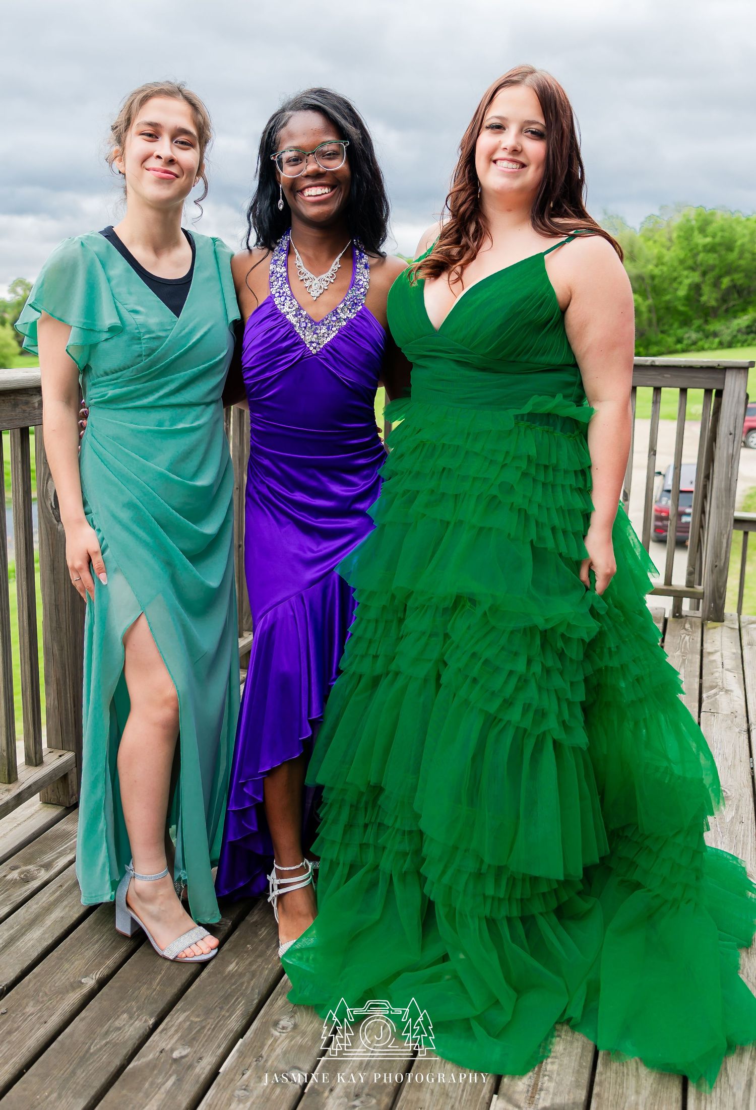 Three friends pose together on a wooden deck wearing elegant formal dresses in green and purple colors.