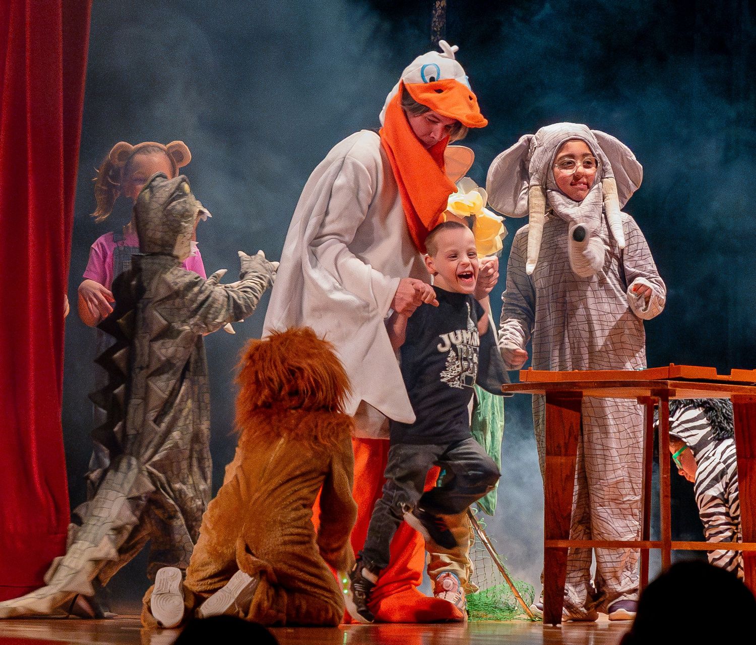 Theatrical performance with costumed actors including animal characters and colorful costumes on a smoky stage.