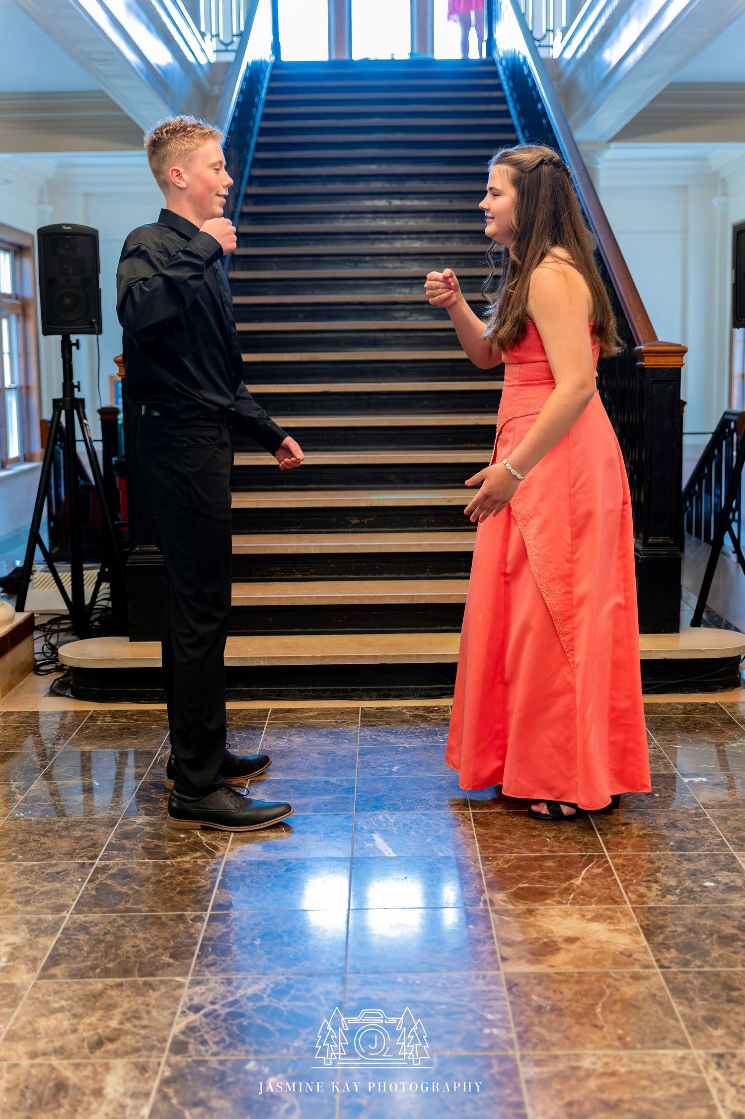 Two people in formal attire share a playful moment on a grand marble staircase in an elegant venue.